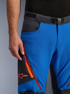 Alpinestars Maxdura Dual Pants w kolorze niebieskim i ciemnoszarym, zbliżenie na pas z klamrą magnetyczną, kieszeń cargo na zamek z pomarańczowymi detalami i czerwonym logo Alpinestars, techniczna odzież motocyklowa adventure