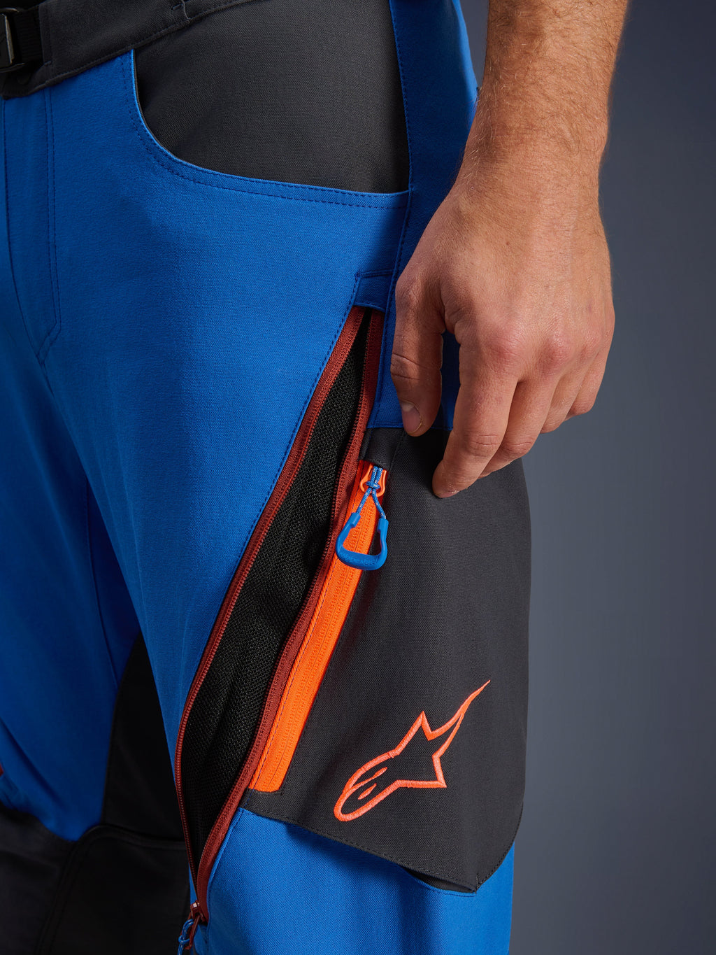 Detale spodni Alpinestars Maxdura Dual, spodnie motocyklowe adventure w kolorze królewskiego błękitu i ciemnoszarym, zbliżenie na zapinaną na zamek wentylację na udzie z pomarańczowymi akcentami i niebieskim uchwytem, pomarańczowe logo Alpinestars na czarnym panelu, wytrzymała konstrukcja z elastycznego poliamidu