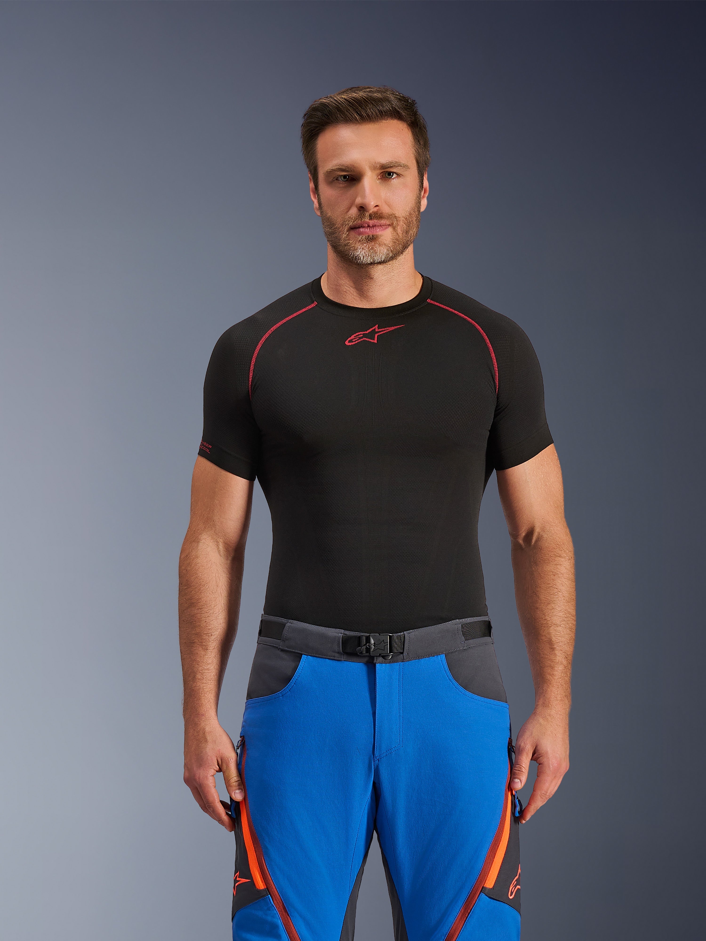 Alpinestars Maxdura Dual Pants, niebieskie i ciemnoszare, spodnie motocyklowe adventure z pomarańczowymi akcentami, noszone przez modela w czarnej koszulce kompresyjnej, pasek z klamrą magnetyczną i zapinane na zamek otwory wentylacyjne na udach, profesjonalna odzież motorsportowa
