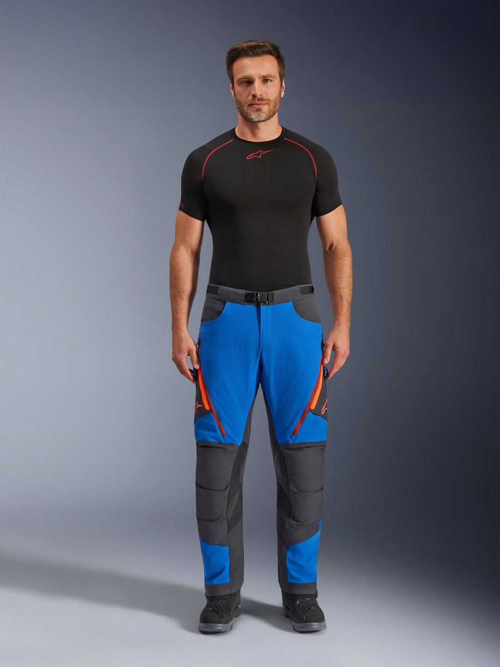 Spodnie Alpinestars Maxdura Dual, motocyklowe spodnie adventure, niebieskie i ciemnoszare z pomarańczowymi akcentami przy zamkach, zestawione z czarną warstwą bazową Alpinestars i czarnymi butami, wzmocnione panele na kolanach i techniczny design off-road