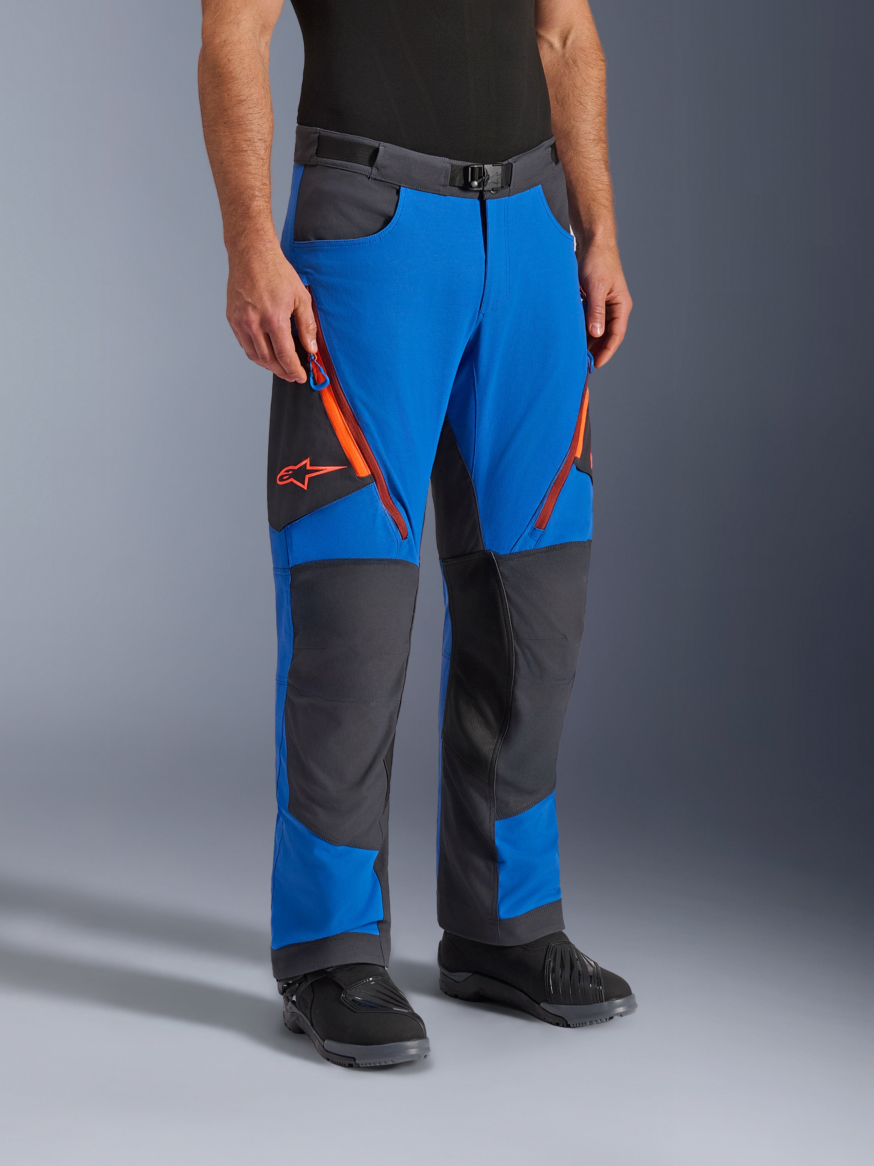 Alpinestars Maxdura Dual Pants, motocyklowe spodnie adventure, kolor Royal Blue i Deep Gray z pomarańczowymi akcentami, z wentylacyjnymi zamkami na udach, wzmocnionymi panelami na kolanach i paskiem z klamrą magnetyczną, techniczny krój na buty do jazdy dual-sport.