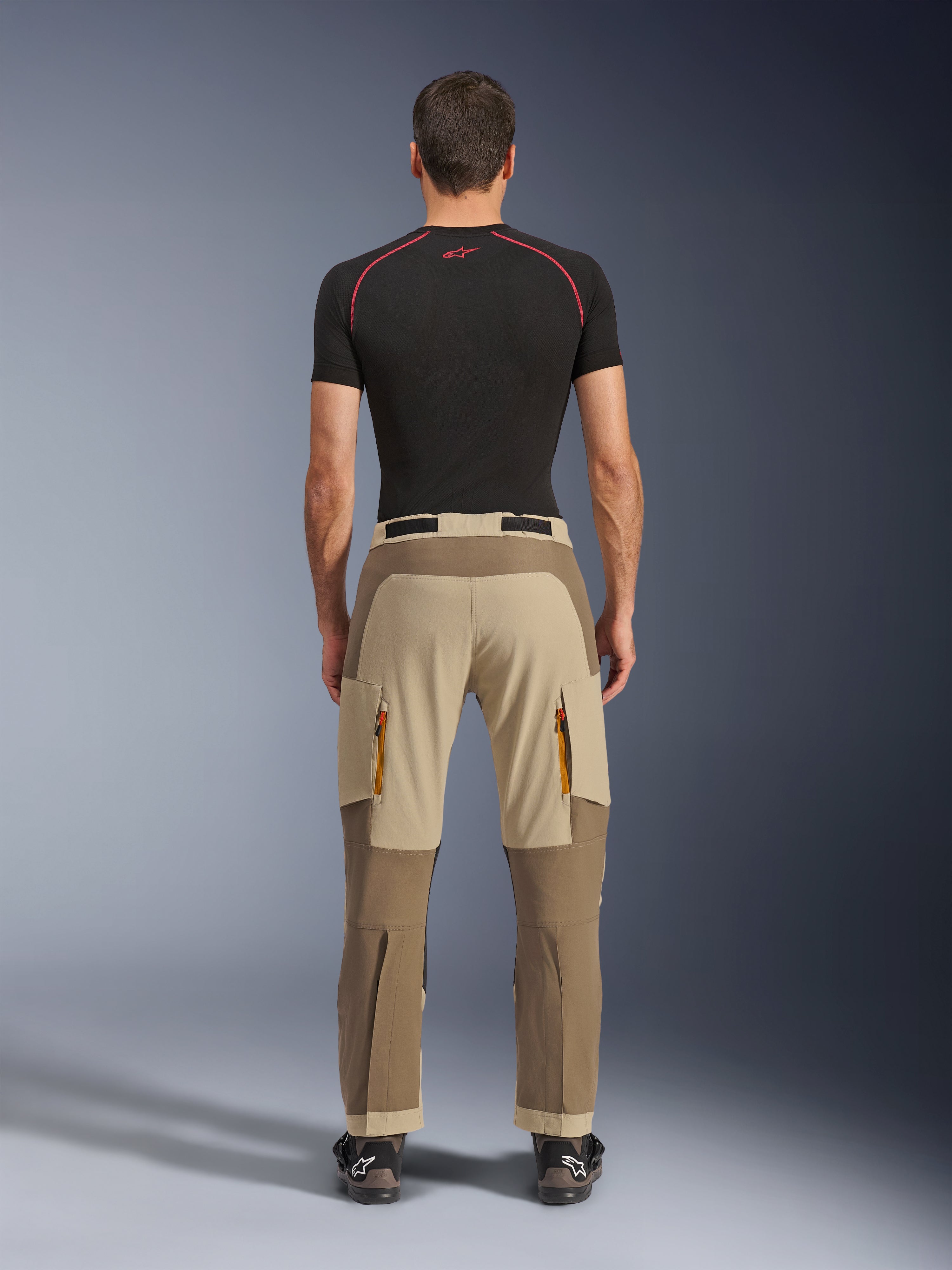 Alpinestars Maxdura Dual Pants in Walnut Aluminum, rear view showing two-tone brown and tan design, adventure riding pants with zippered cargo pockets and reinforced seat, worn with a black compression shirt and motorcycling boots.