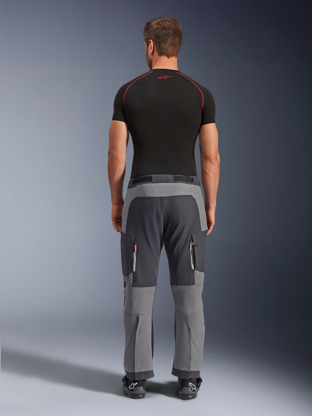 Spodnie Alpinestars Maxdura Dual, motocyklowe spodnie adventure w kolorach Dark Gray i Deep Gray, widok z tyłu na modelu w czarnym t-shircie i butach, z szarymi panelami o różnych odcieniach i kieszeniami cargo na zamek z czerwonymi akcentami.