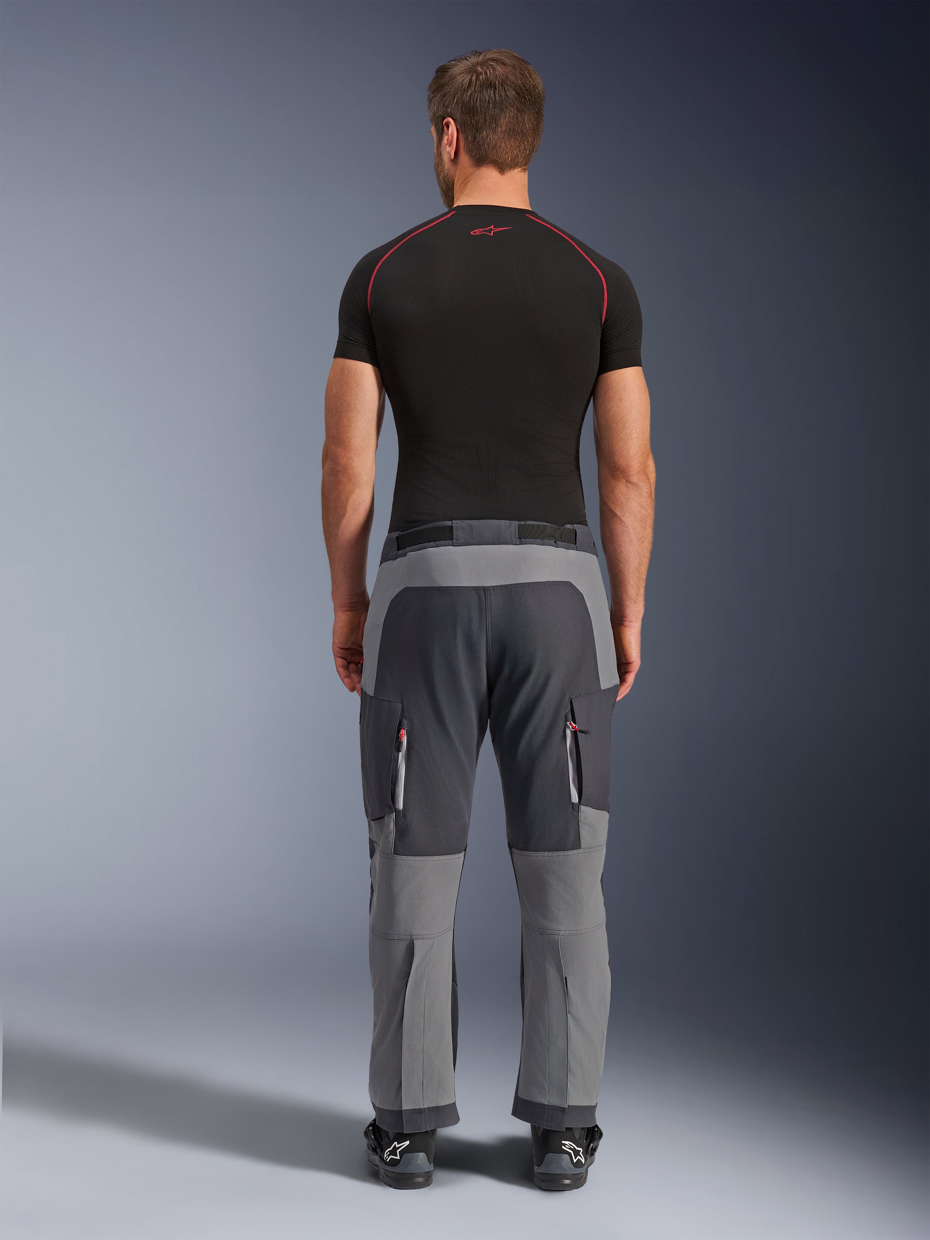 Spodnie Alpinestars Maxdura Dual, motocyklowe spodnie adventure w kolorach Dark Gray i Deep Gray, widok z tyłu na modelu w czarnym t-shircie i butach, z szarymi panelami o różnych odcieniach i kieszeniami cargo na zamek z czerwonymi akcentami.