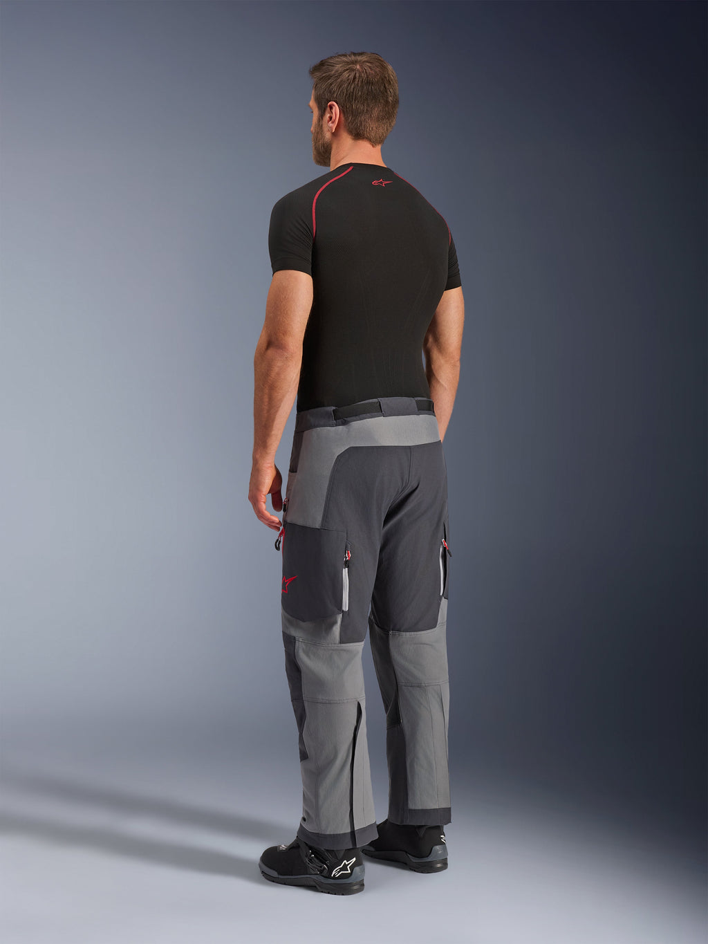 Spodnie Alpinestars Maxdura Dual, spodnie motocyklowe adventure, ciemnoszare i grafitowe, widoczne na modelu od tyłu z boku, czerwone akcenty na zamkach i logo, wzmocnione szare panele, kieszenie cargo, krój nakładany na buty
