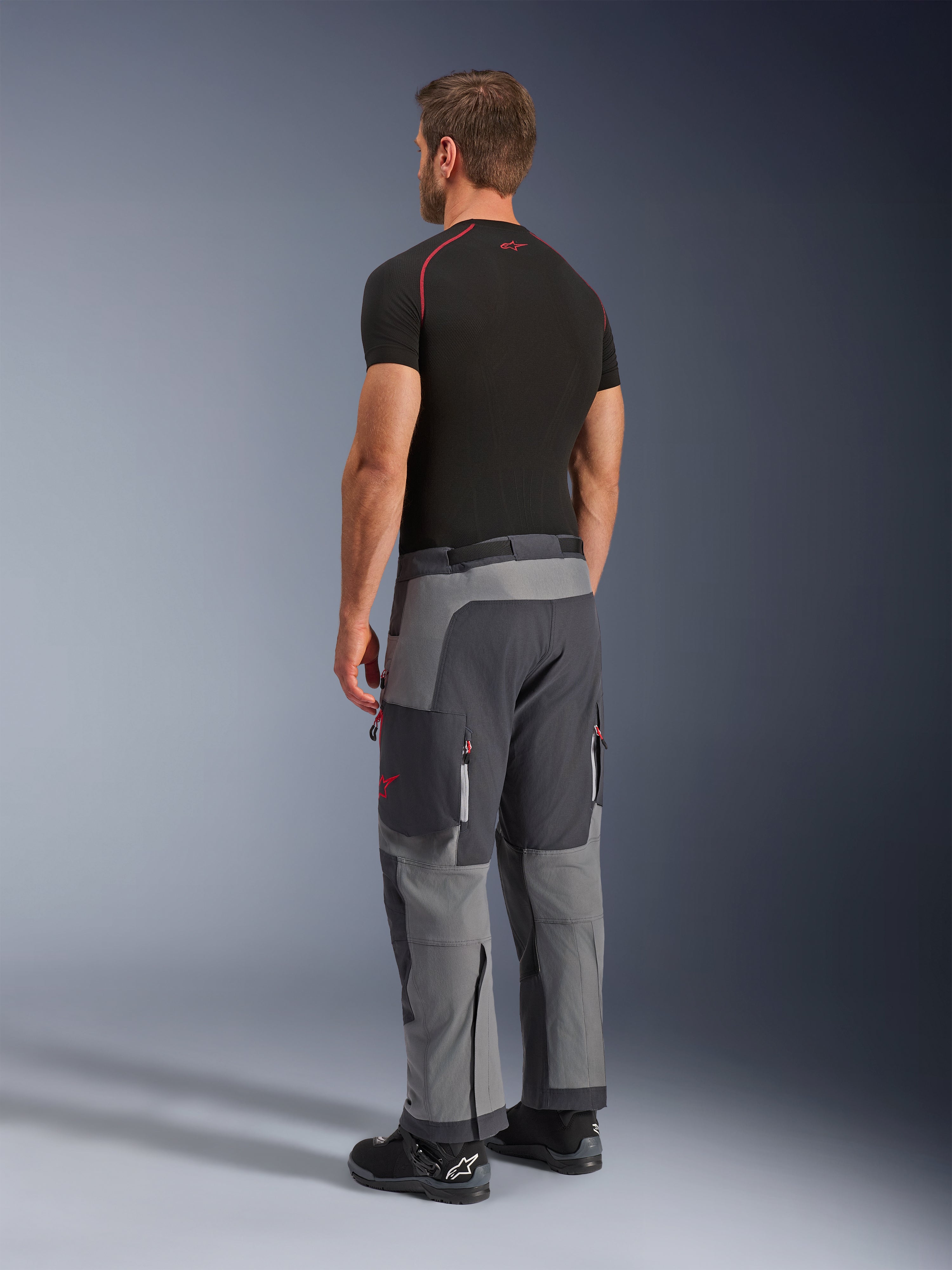 Spodnie Alpinestars Maxdura Dual, spodnie motocyklowe adventure, ciemnoszare i grafitowe, widoczne na modelu od tyłu z boku, czerwone akcenty na zamkach i logo, wzmocnione szare panele, kieszenie cargo, krój nakładany na buty