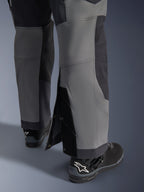 Alpinestars Maxdura Dual Pants, motocyklowe spodnie adventure, ciemnoszare i głęboko szare, zbliżenie na regulowany dół nogawki typu over-the-boot z zapięciami na zatrzaski i wzmocnionymi panelami, noszone z czarnymi butami Alpinestars, wytrzymała techniczna konstrukcja do jazdy dual-sport