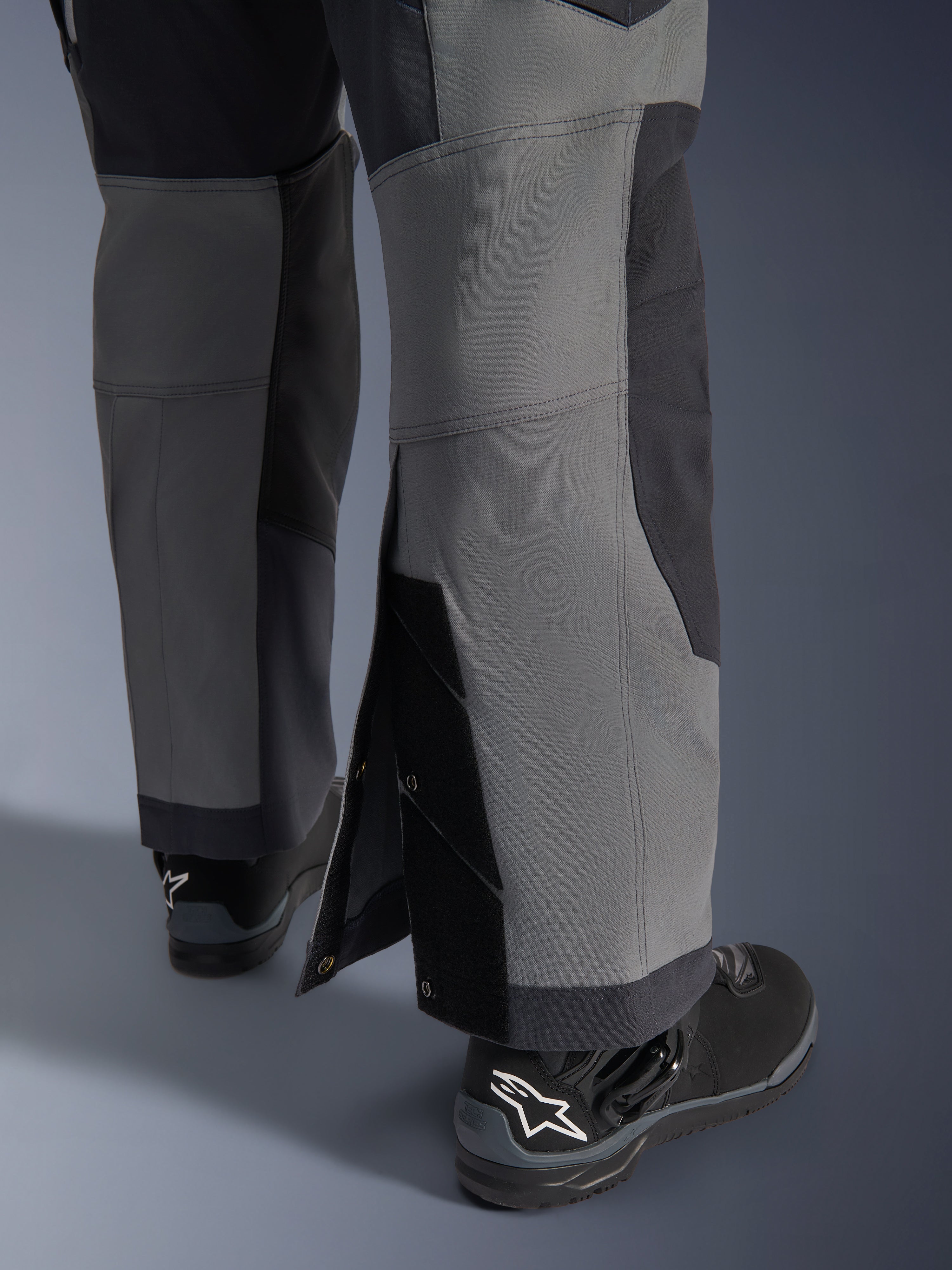Alpinestars Maxdura Dual Pants, motocyklowe spodnie adventure, ciemnoszare i głęboko szare, zbliżenie na regulowany dół nogawki typu over-the-boot z zapięciami na zatrzaski i wzmocnionymi panelami, noszone z czarnymi butami Alpinestars, wytrzymała techniczna konstrukcja do jazdy dual-sport