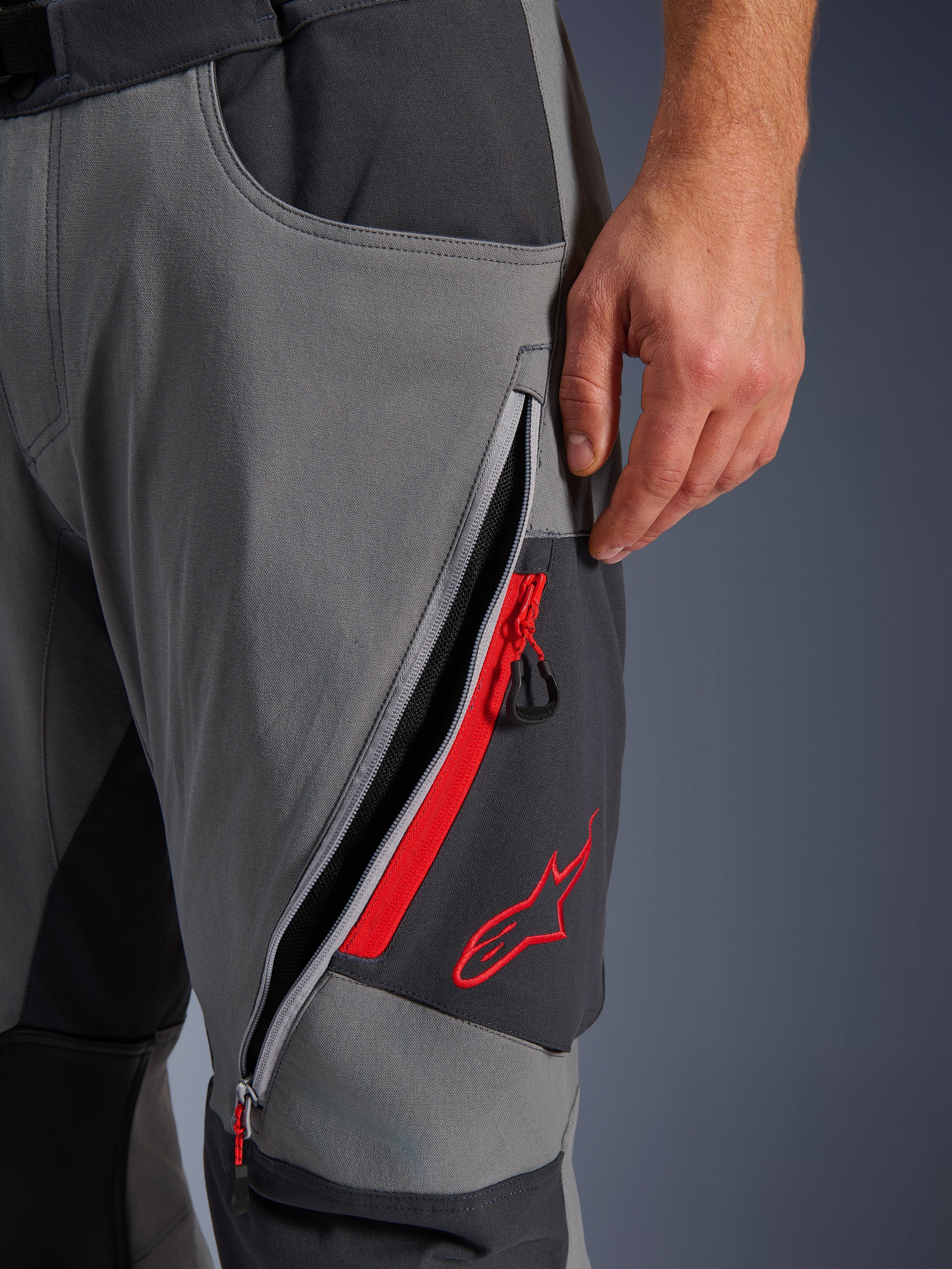 Alpinestars Maxdura Dual Pants, ciemnoszare spodnie motocyklowe adventure, zbliżenie na otwarty otwór wentylacyjny na udzie z czerwonymi akcentami i haftowanym czerwonym logo gwiazdy, techniczna elastyczna tkanina