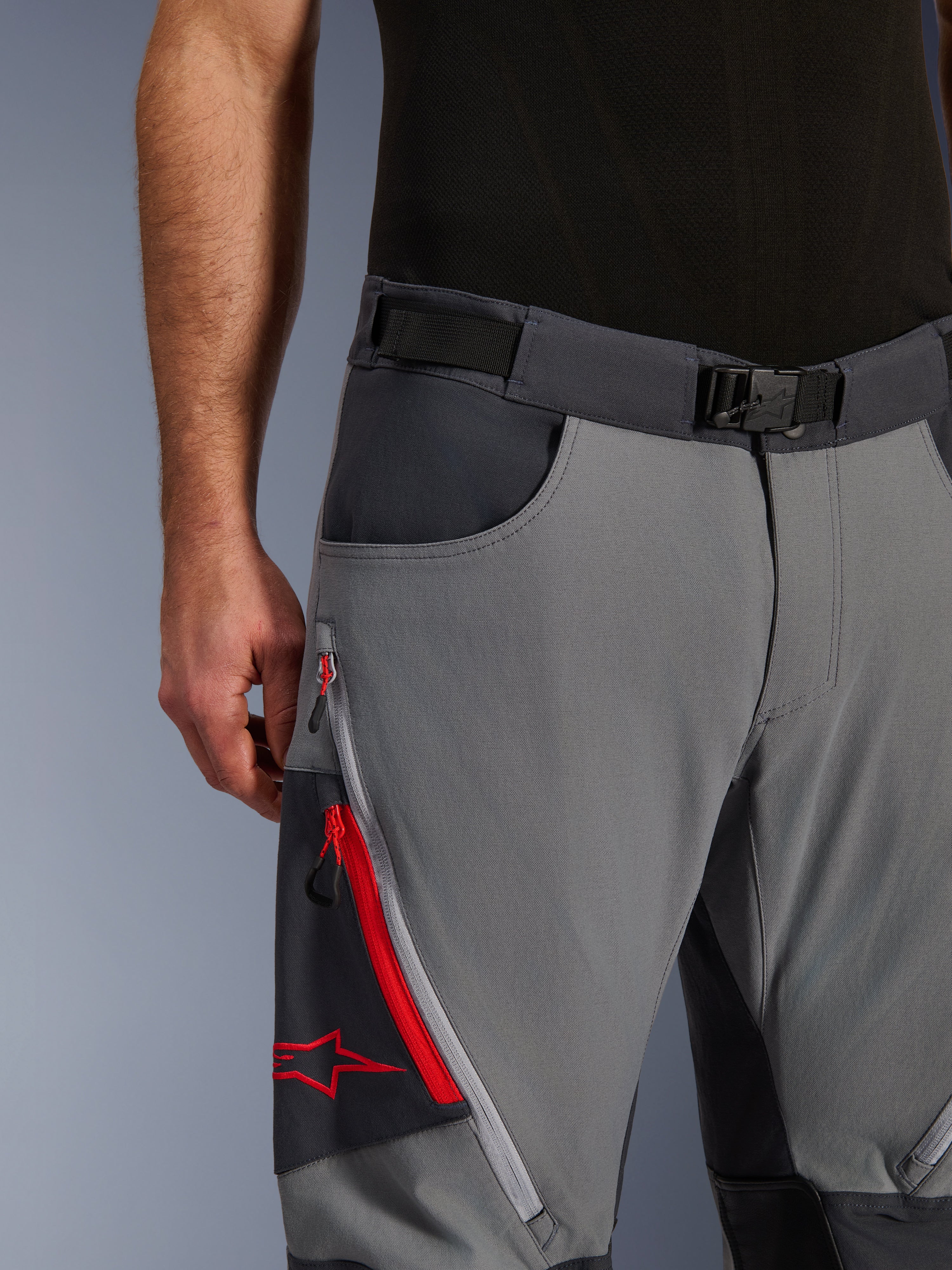 Alpinestars Maxdura Dual Pants, spodnie motocyklowe adventure, ciemnoszare i głęboko szare, zbliżenie na regulowany pasek w talii z klamrą magnetyczną, kieszeń cargo na zamek z czerwonymi detalami i logo Alpinestars