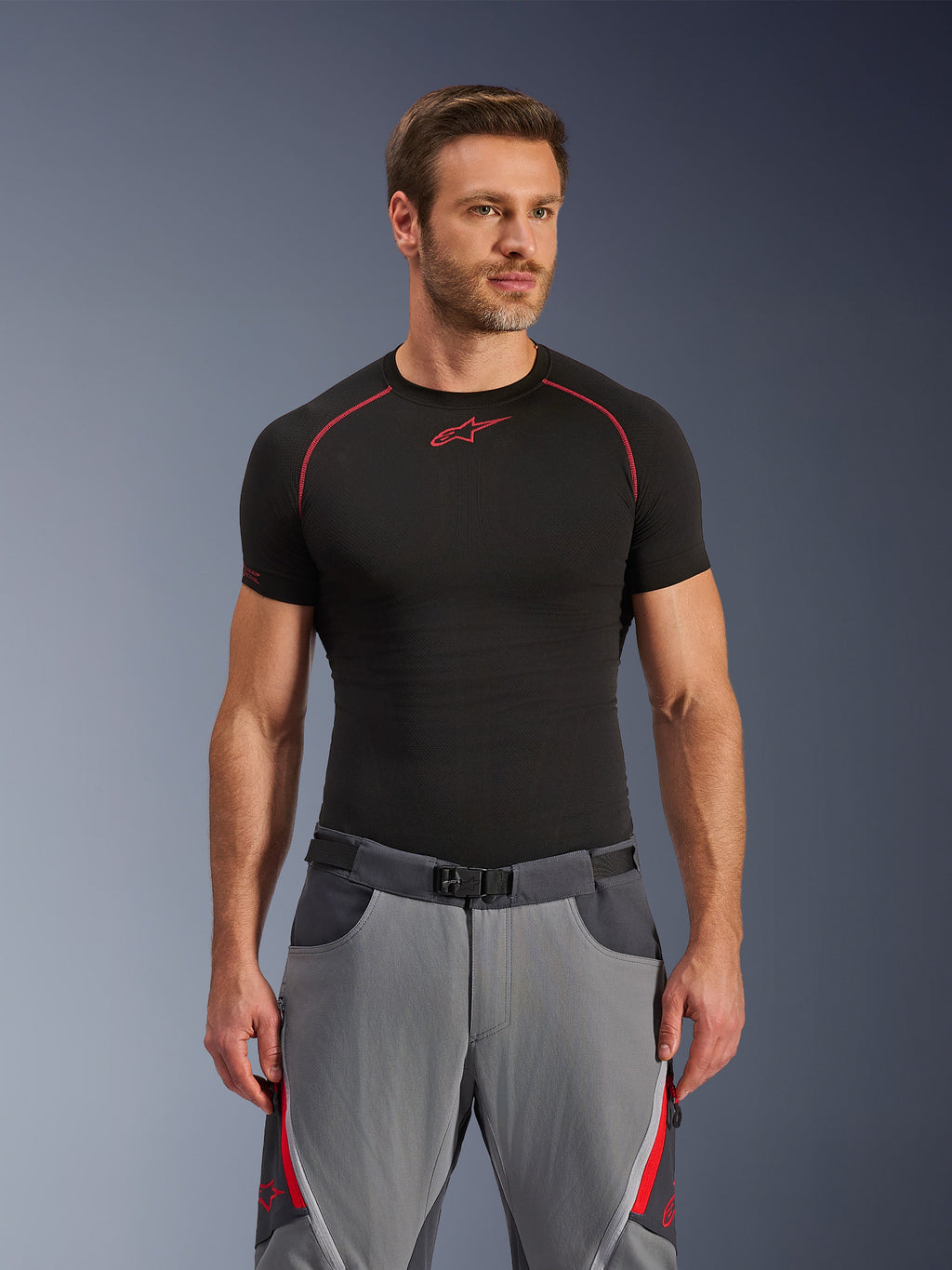 Alpinestars Maxdura Dual Pants, ciemnoszare i głęboko szare z czerwonymi akcentami, noszone z czarną koszulką termoaktywną z czerwonymi przeszyciami, sportowy krój ze zintegrowanym paskiem z magnetyczną klamrą, wytrzymała odzież motocyklowa adventure