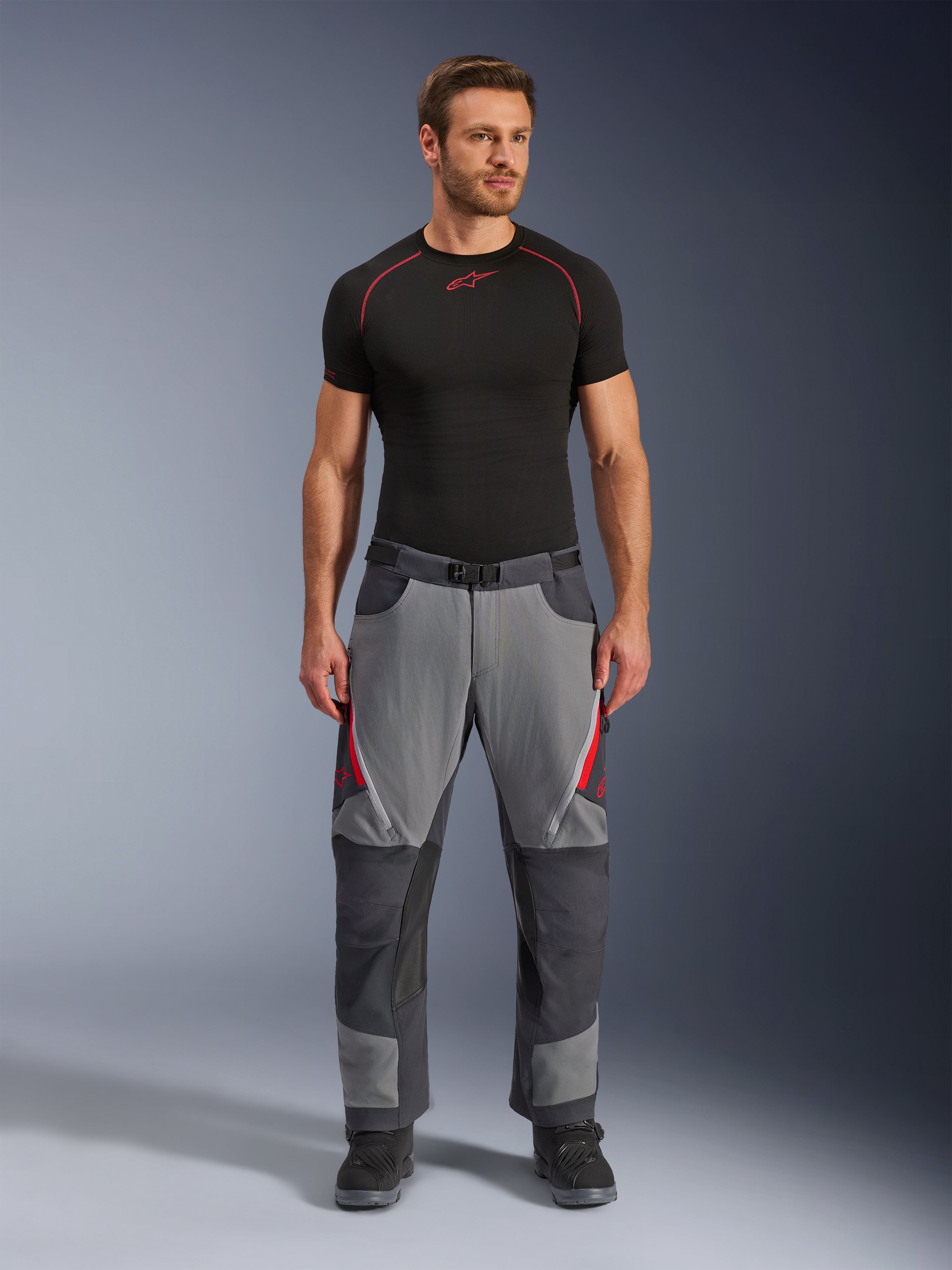 Spodnie motocyklowe Alpinestars Maxdura Dual, ciemnoszare i szare z czerwonymi akcentami, widok z przodu na modelu w czarnej warstwie bazowej Alpinestars i czarnych butach, wzmocnione panele i zapinane na zamek otwory wentylacyjne na udach, odzież motocyklowa typu adventure