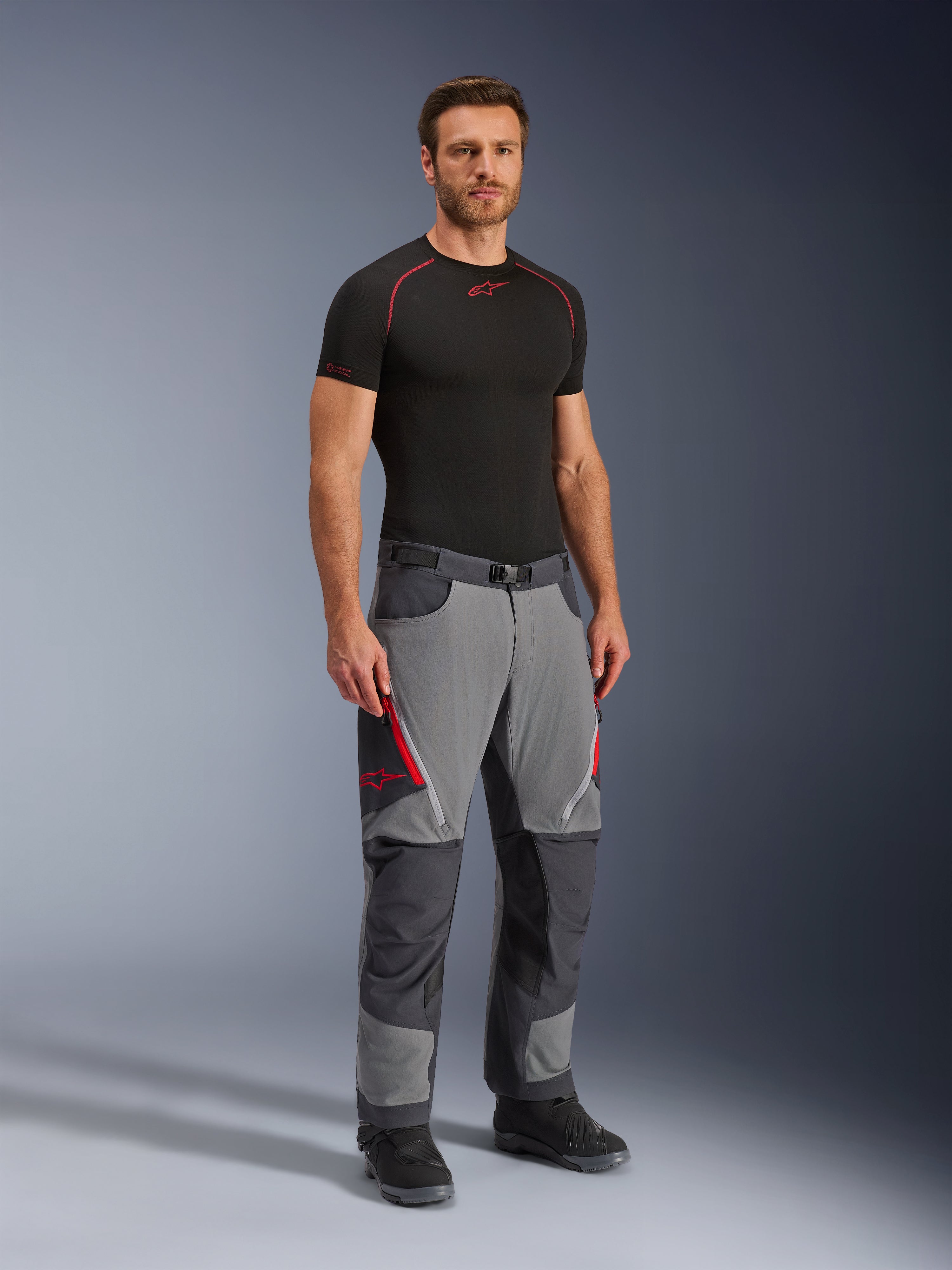 Alpinestars Maxdura Dual Pants, spodnie motocyklowe adventure, ciemnoszare i grafitowe, noszone z czarną warstwą bazową i butami, z czerwonymi otworami wentylacyjnymi na zamki, wzmocnionymi panelami na kolanach i siedzeniu oraz paskiem w talii, sportowy krój do jazdy dual-sport