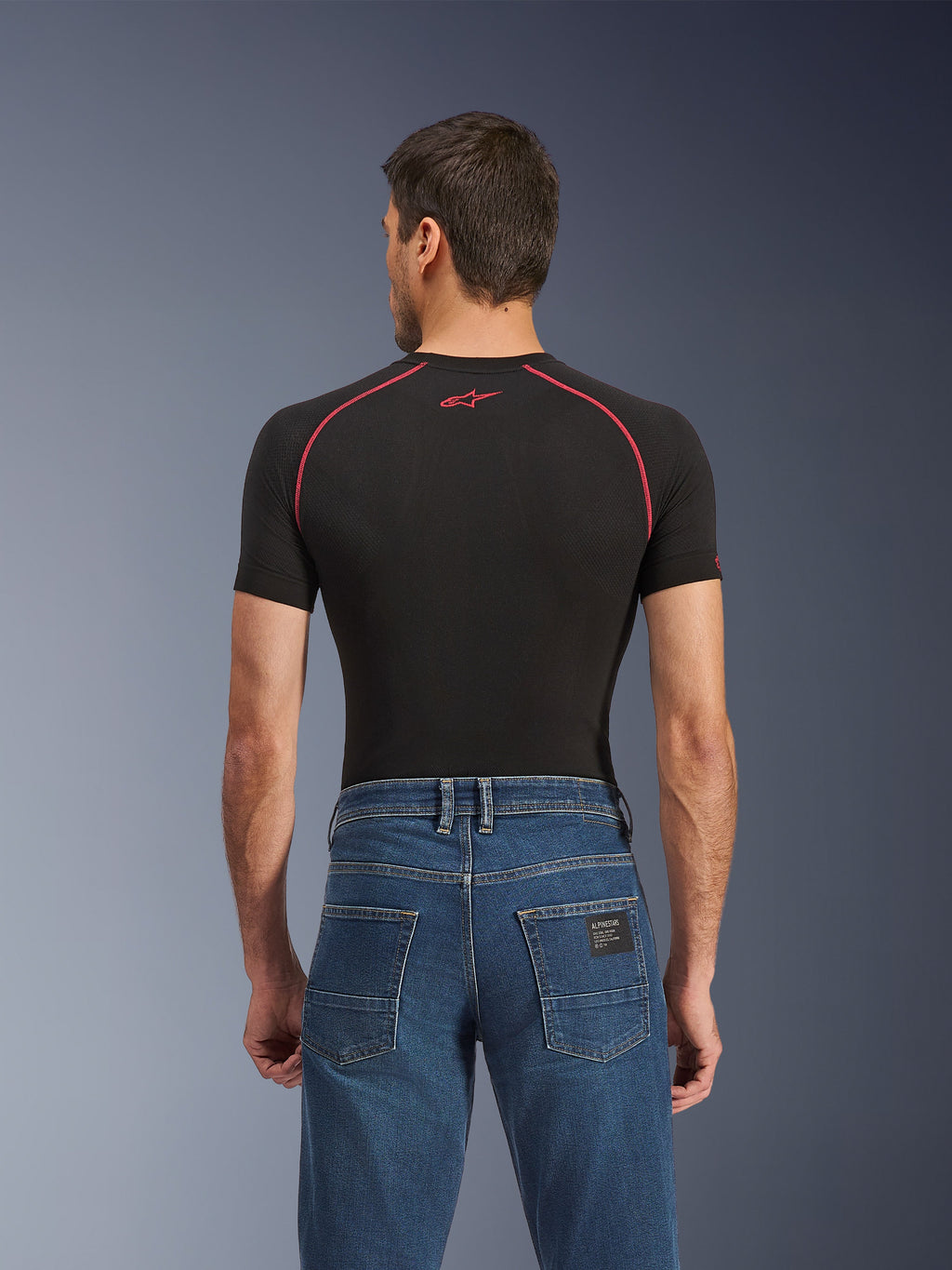 Spodnie jeansowe Alpinestars Flex-AST 5, Indigo Washed, widok z tyłu pokazujący regularny zwężany krój, noszone z czarną warstwą bazową z krótkim rękawem i czerwonymi przeszyciami, codzienne miejskie jeansy motocyklowe.