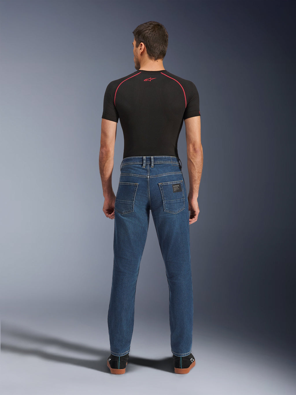 Mężczyzna widziany od tyłu ubrany w spodnie Alpinestars Flex-AST 5 Denim w kolorze Indigo Washed, miejskie jeansy motocyklowe o nowoczesnym, zwężanym kroju i tylnych kieszeniach, zestawione z czarną warstwą bazową z krótkim rękawem i codziennymi czarnymi butami.