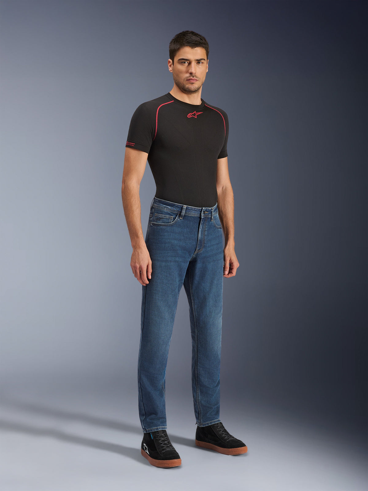 Alpinestars Flex-AST 5 Denim Pants - Short, Indigo Washed, miejskie dżinsy motocyklowe o zwężanym kroju, prezentowane przez modela w czarnej dopasowanej koszulce i czarnych sneakersach, klasyczny styl dżinsowy zapewniający płynne przejście z drogi na ulicę