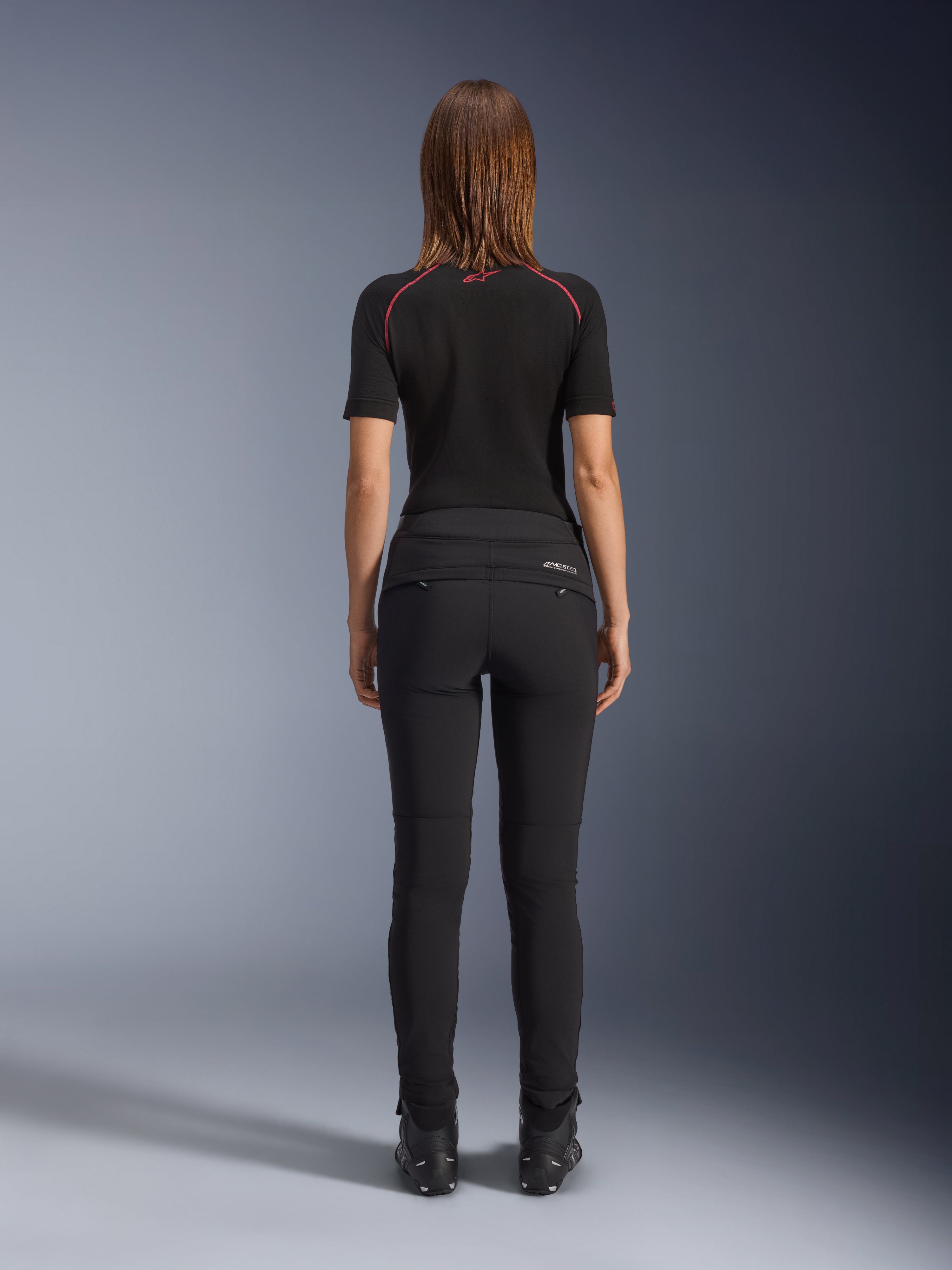 Alpinestars Alamosa Women Pants, czarne, widok z tyłu prezentujący dopasowaną konstrukcję tekstylną, tylne kieszenie zapinane na zamek oraz odblaskowe logo MO.ST.EQ. na karczku, zestawione z czarną warstwą bazową z krótkim rękawem i czarnymi butami motocyklowymi