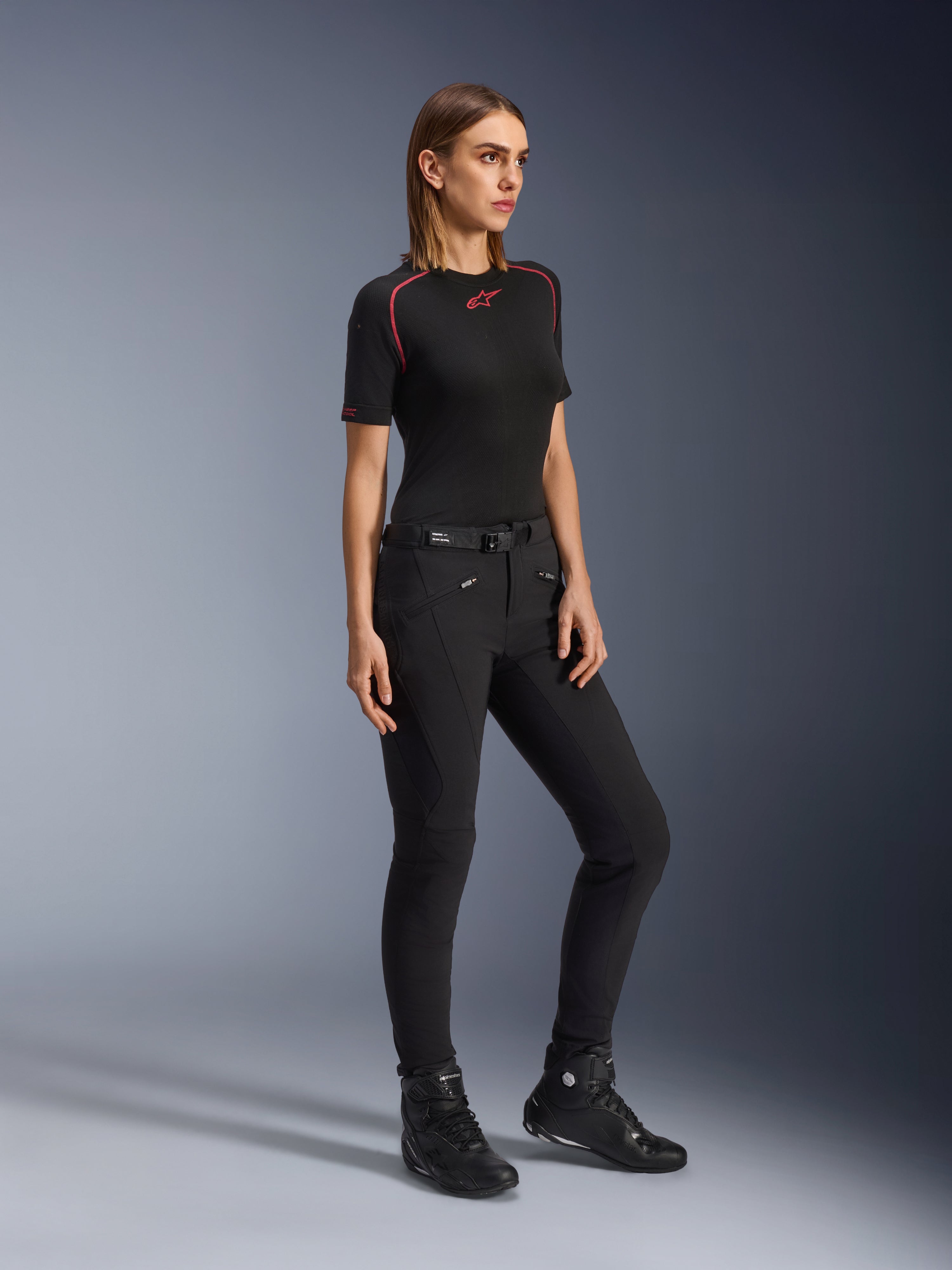 Alpinestars Alamosa Women Pants, damskie spodnie motocyklowe o kroju slim-fit w kolorze czarnym, prezentowane przez modelkę w czarnej warstwie bazowej i butach motocyklowych, wyposażone w przednie kieszenie na zamek i pasek z magnetycznym zapięciem, miejski styl streetwear.