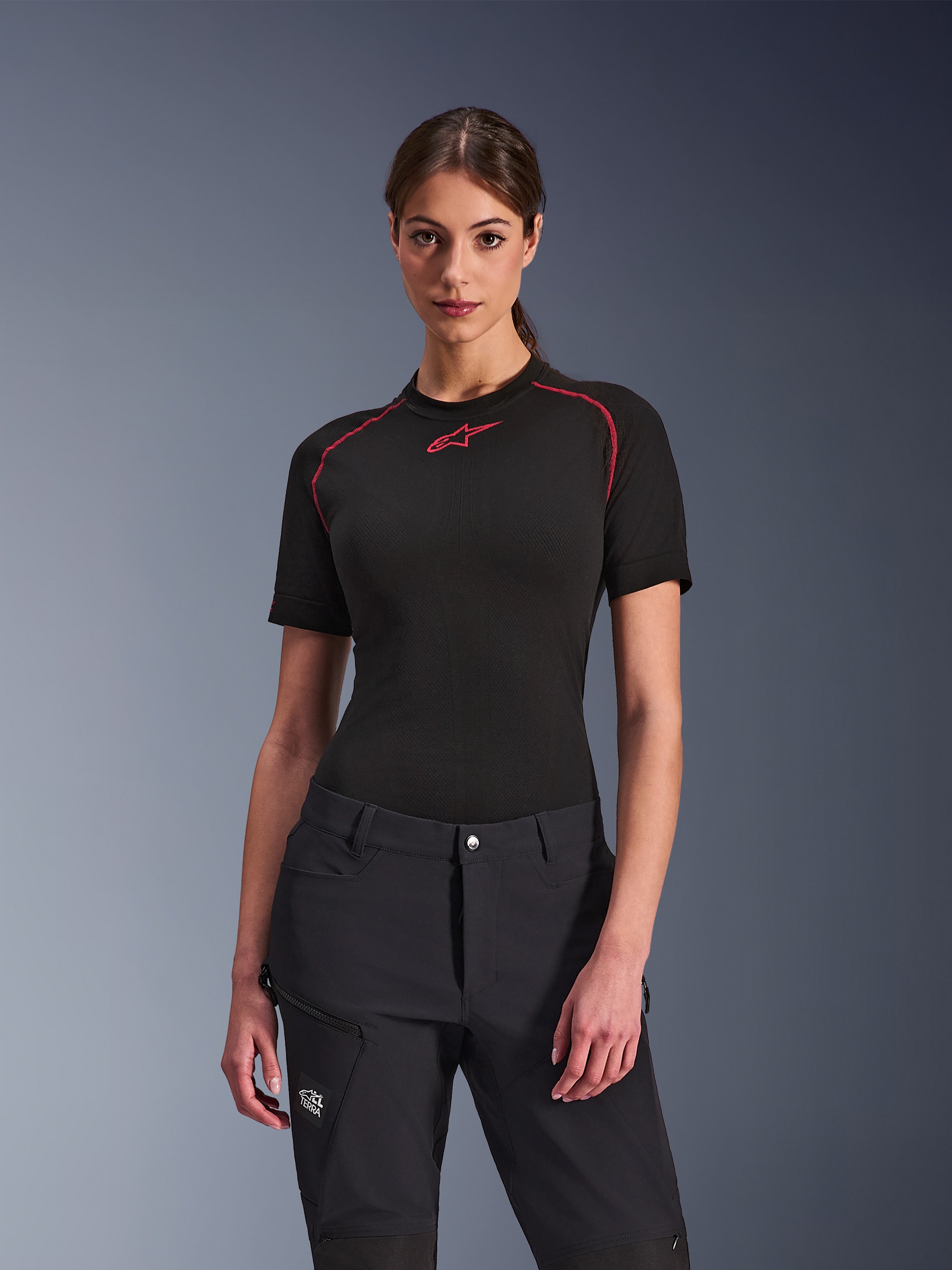 Kobieta ubrana w czarne spodnie Alpinestars Stella Flex-AST Explorer, czarna warstwa bazowa z krótkim rękawem i czerwonymi akcentami, odzież motocyklowa adventure z kieszeniami cargo i logo All Terra
