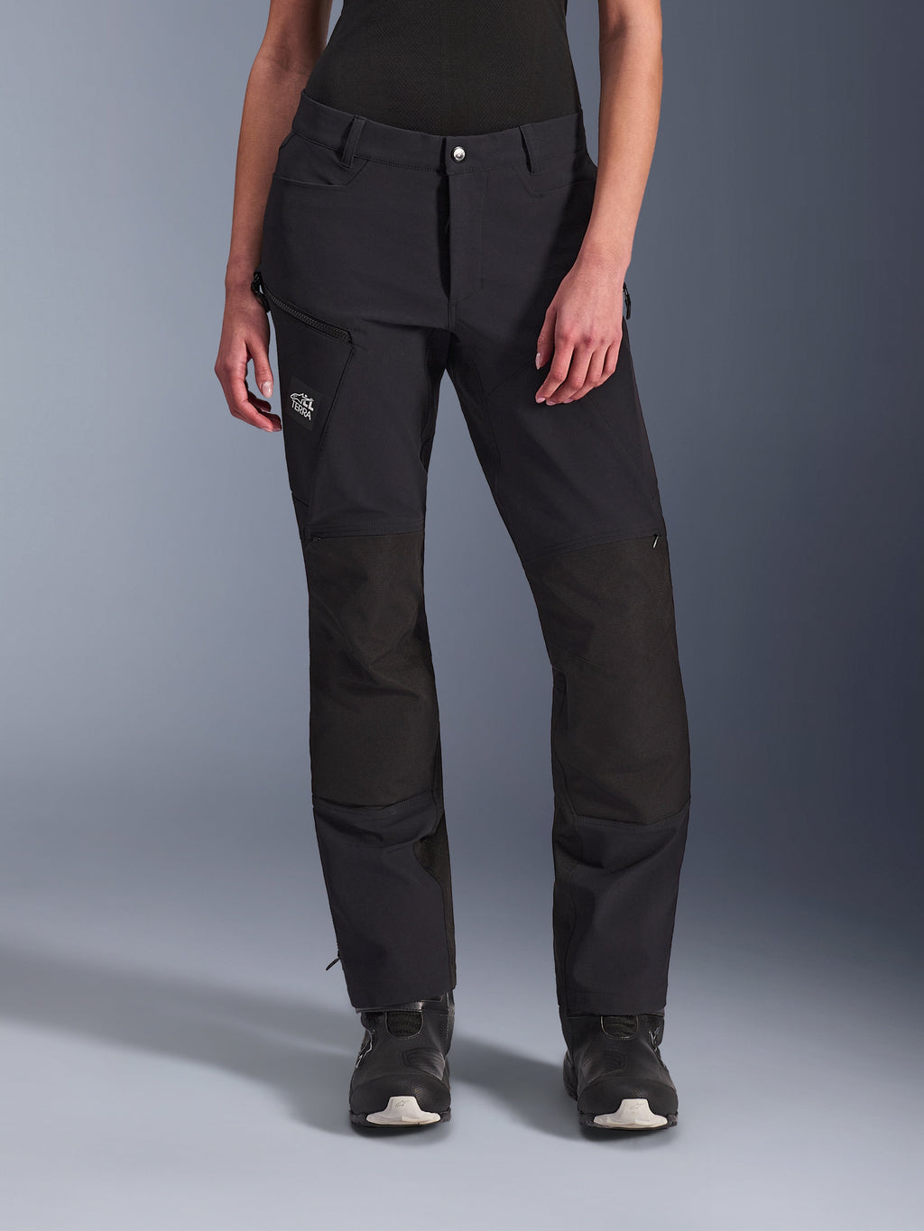 Alpinestars Stella Explore AST Pants, damskie spodnie motocyklowe adventure, czarne, wzmocnienia ripstop na kolanach, kieszeń cargo na udzie zapinana na zamek, logo „Terra”, noszone z czarnymi butami, opływowy design do turystyki przygodowej i jazdy szosowej