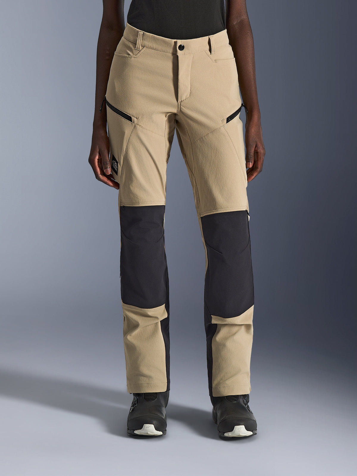 Alpinestars Stella Flex-AST Explorer Pants, damskie spodnie motocyklowe, Aluminum Black, widok z przodu na modelce, piaskowo-beżowa tkanina z czarnymi panelami ripstop na kolanach i dole nogawek, kieszenie na zamek, styl adventure touring