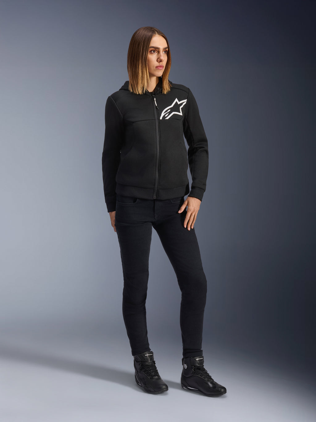 Kobieta ubrana w damskie spodnie motocyklowe Alpinestars Daisy V3 Women's Riding Denim o kroju slim-fit w kolorze Rinse Black, czarna rozpinana bluza z kapturem i buty motocyklowe, miejska casualowa odzież ochronna