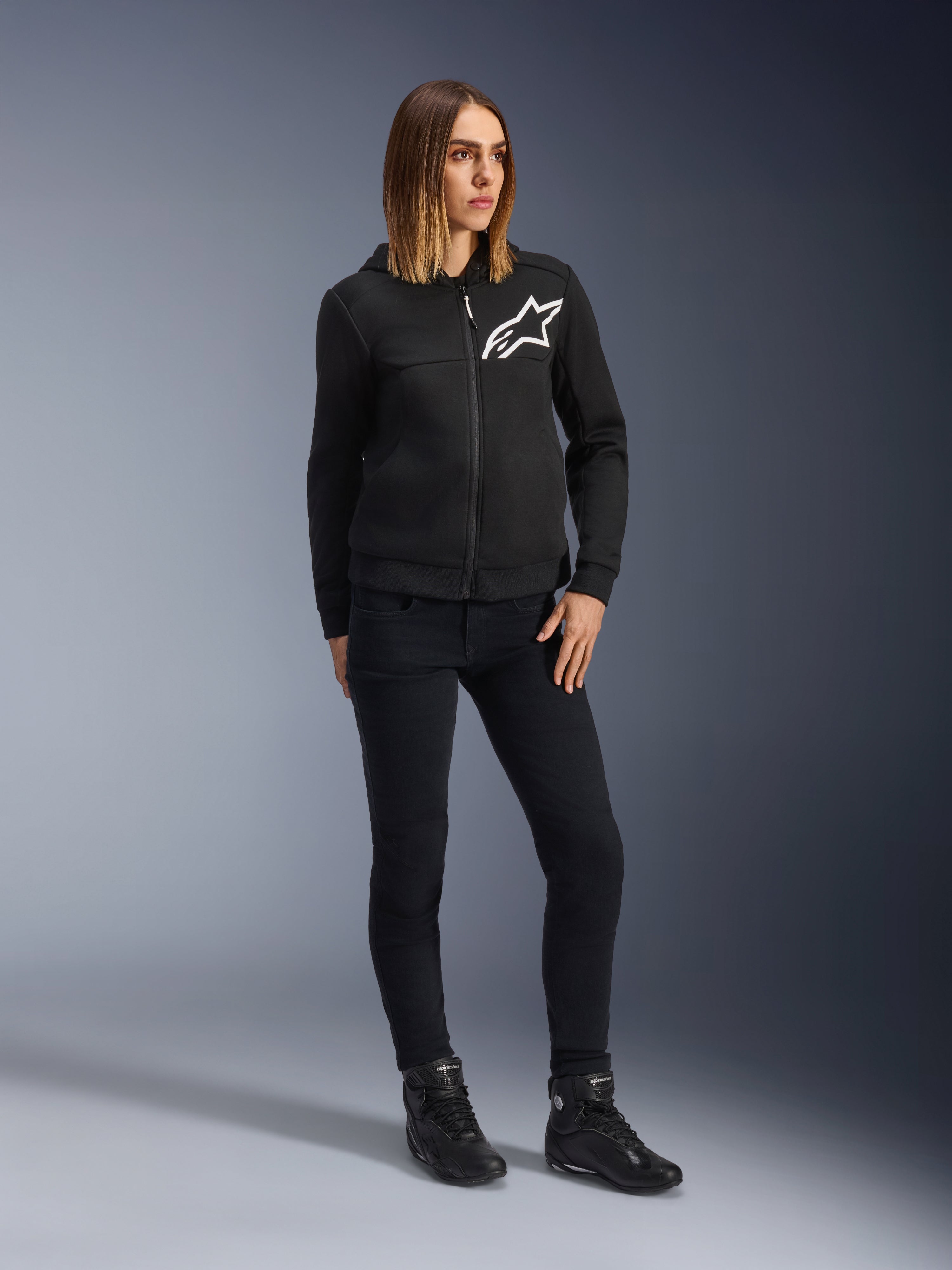 Kobieta ubrana w damskie spodnie motocyklowe Alpinestars Daisy V3 Women's Riding Denim o kroju slim-fit w kolorze Rinse Black, czarna rozpinana bluza z kapturem i buty motocyklowe, miejska casualowa odzież ochronna