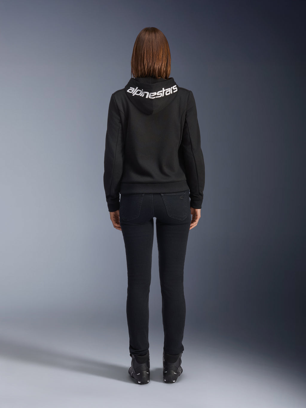 Jeansy Daisy V3 Damskie, Black Rinse, spodnie motocyklowe slim fit, widok z tyłu pokazujący podwyższony stan i kobiecą sylwetkę, zestawione z czarną bluzą z kapturem Alpinestars i czarnymi butami motocyklowymi, miejska odzież casual