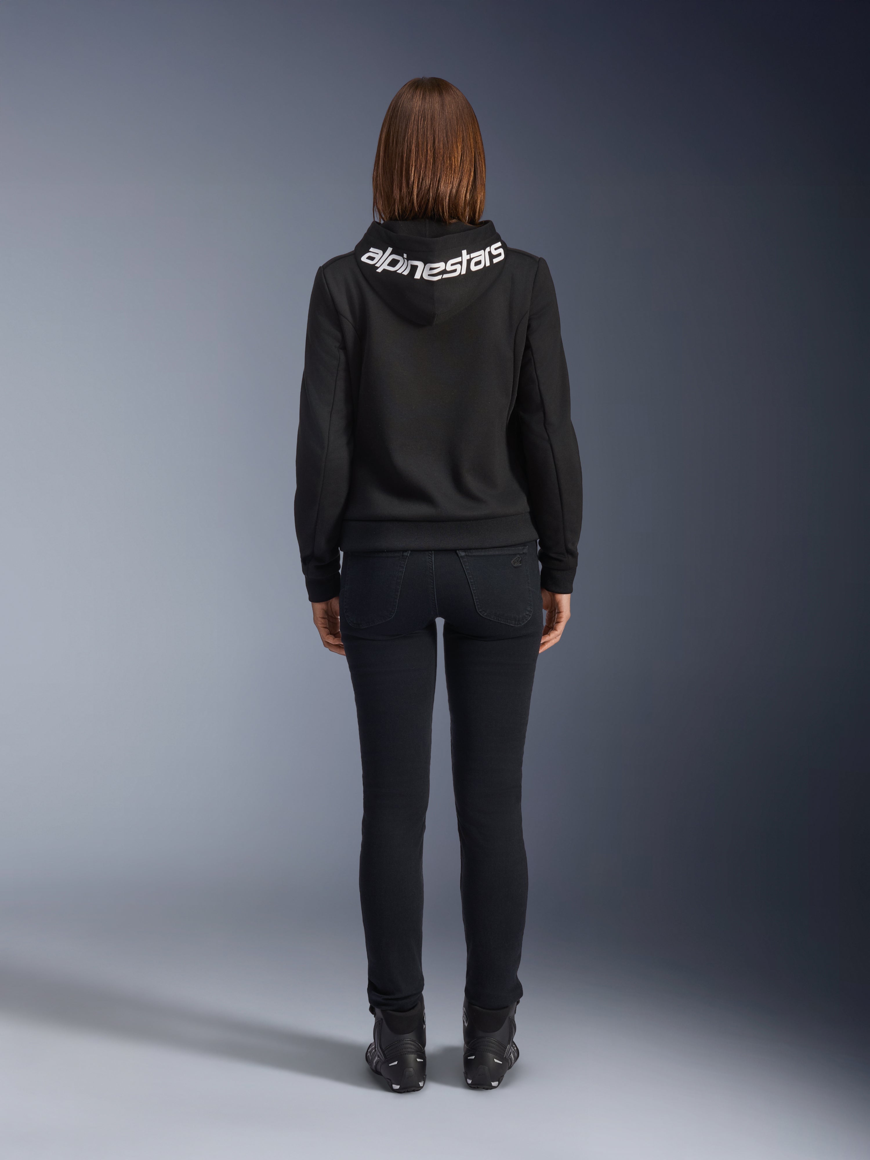 Jeansy Daisy V3 Damskie, Black Rinse, spodnie motocyklowe slim fit, widok z tyłu pokazujący podwyższony stan i kobiecą sylwetkę, zestawione z czarną bluzą z kapturem Alpinestars i czarnymi butami motocyklowymi, miejska odzież casual