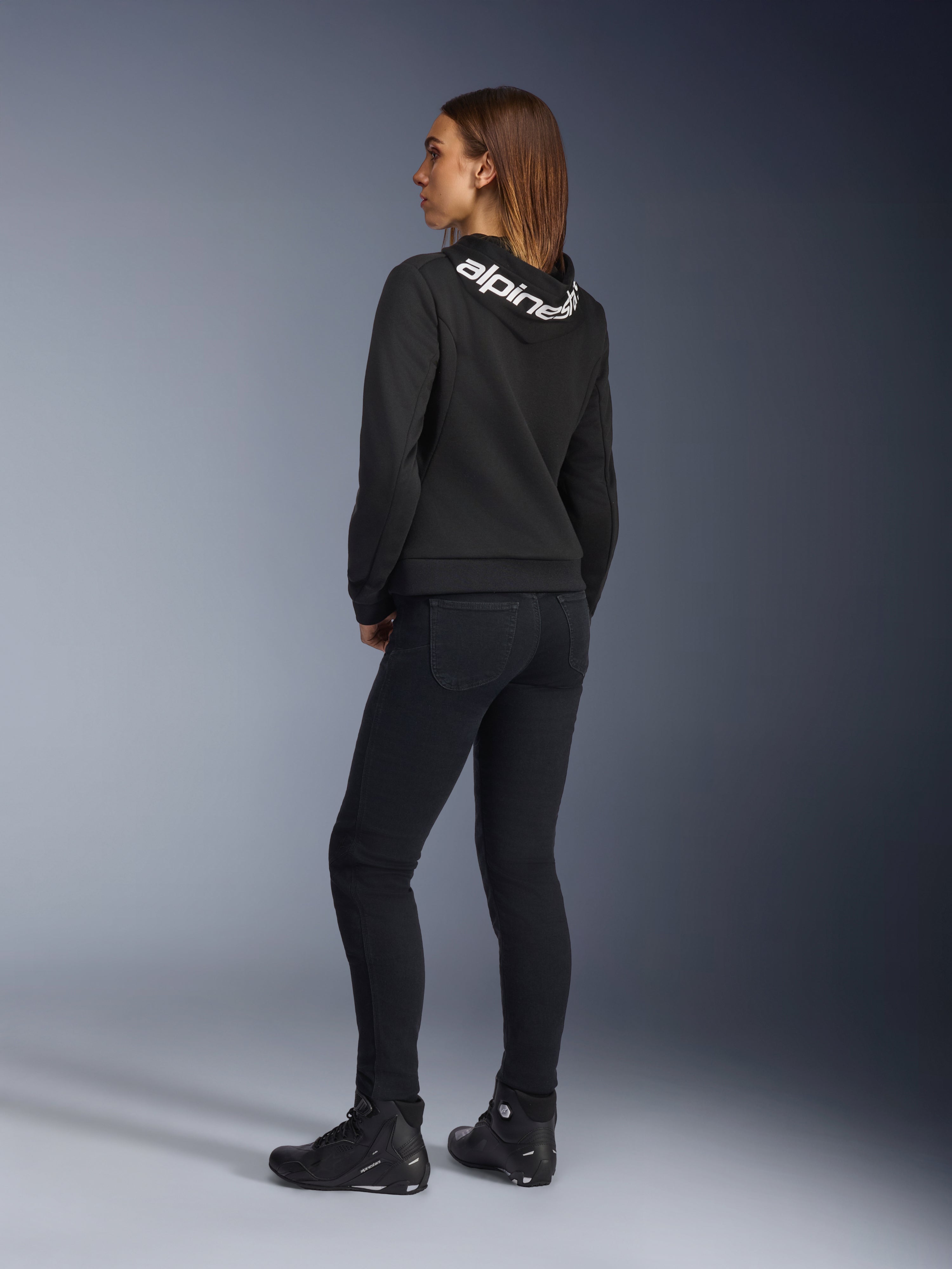 Alpinestars Daisy V3 Women's Riding Denim, damskie spodnie motocyklowe, Rinse Black, widok z tyłu prezentujący dopasowany krój slim fit i podwyższony stan z tyłu, w zestawieniu z czarną bluzą z kapturem Alpinestars i butami motocyklowymi w miejskim stylu
