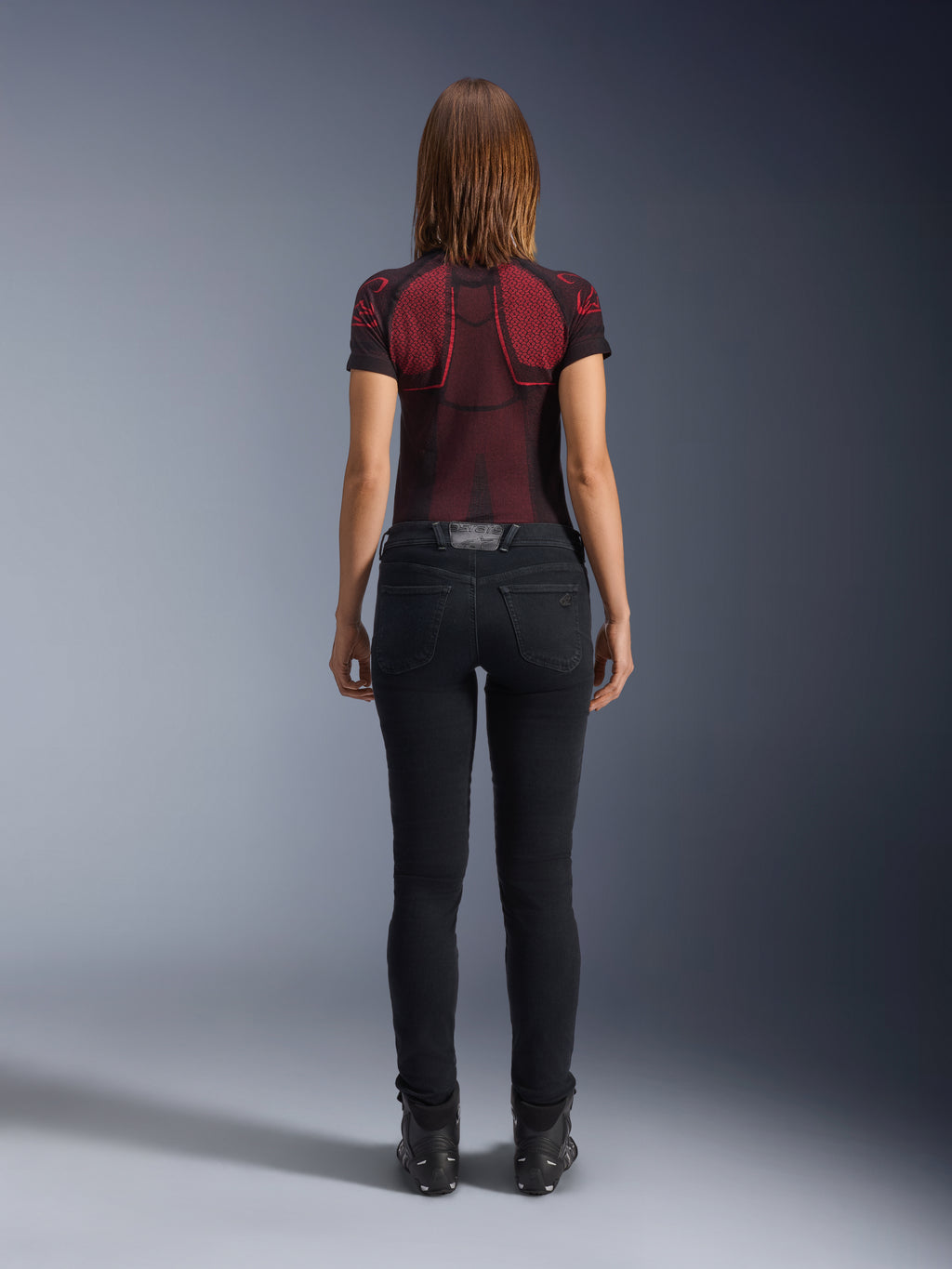 Alpinestars Daisy V3 Women's Riding Denim, Rinse Black, damskie spodnie motocyklowe o kroju slim fit widoczne od tyłu, wysoki stan i tylne kieszenie, w zestawieniu z czerwono-czarną wzorzystą koszulką techniczną i czarnymi butami, miejska odzież motocyklowa dla kobiet