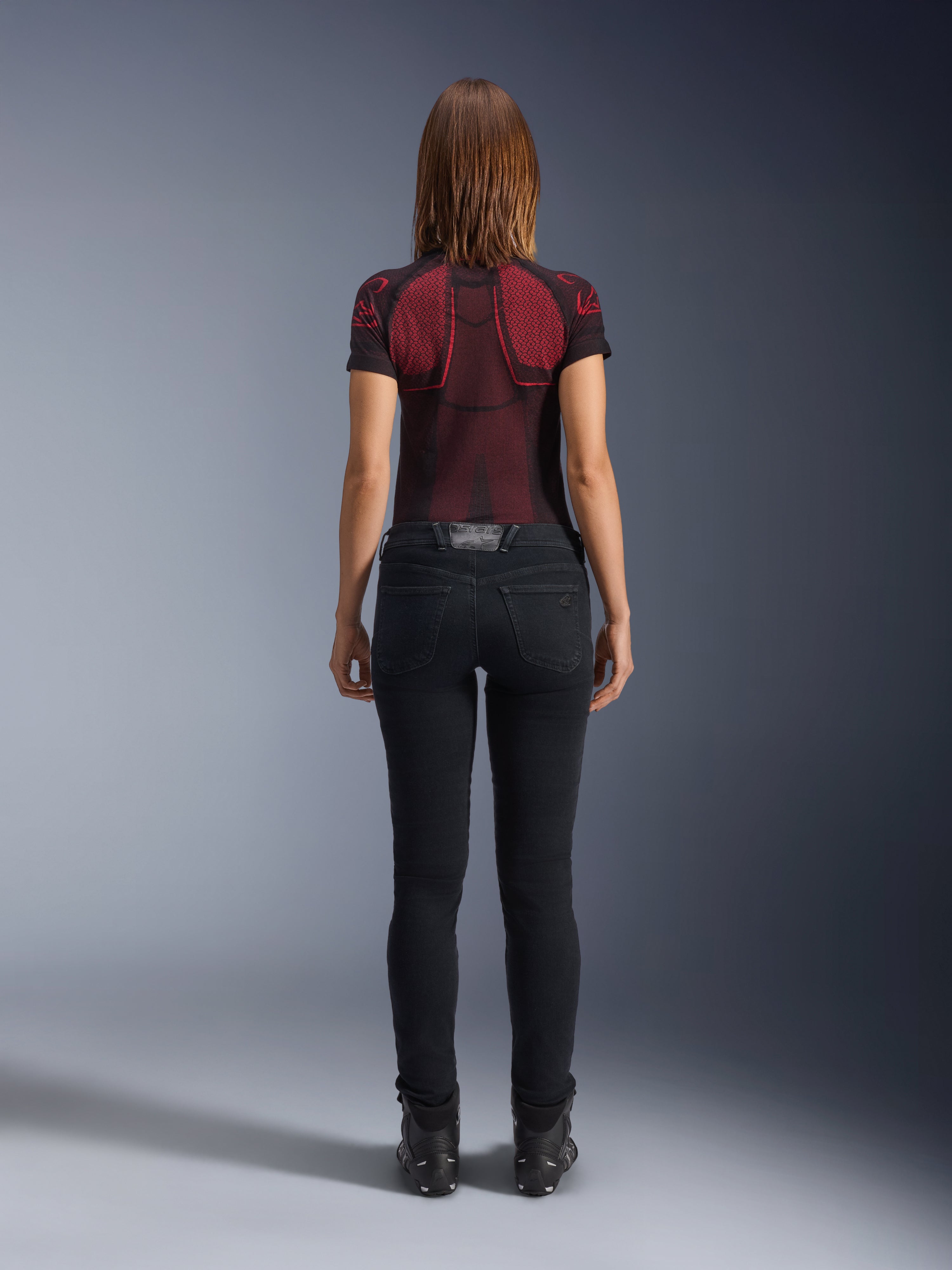 Alpinestars Daisy V3 Women's Riding Denim, Rinse Black, damskie spodnie motocyklowe o kroju slim fit widoczne od tyłu, wysoki stan i tylne kieszenie, w zestawieniu z czerwono-czarną wzorzystą koszulką techniczną i czarnymi butami, miejska odzież motocyklowa dla kobiet
