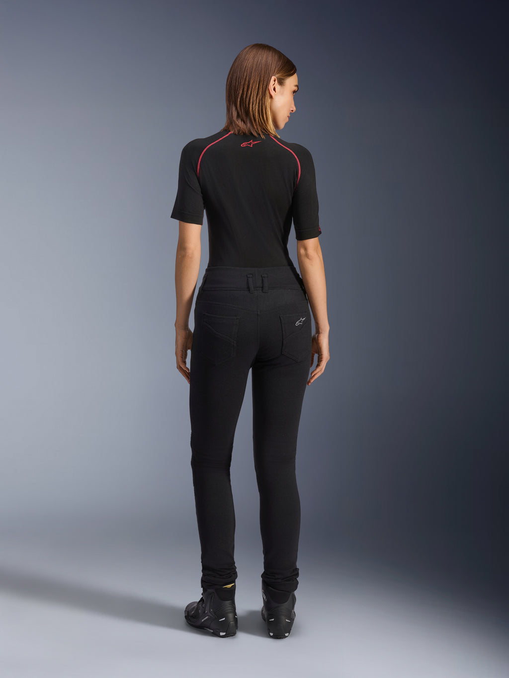 Damskie legginsy Alpinestars Banshee, czarne legginsy motocyklowe, widok z tyłu prezentujący dopasowany krój i anatomiczną elastyczną tkaninę, z tylnymi kieszeniami i detalami z logo, zestawione z czarną warstwą bazową i butami motocyklowymi, miejska odzież motocyklowa dla kobiet