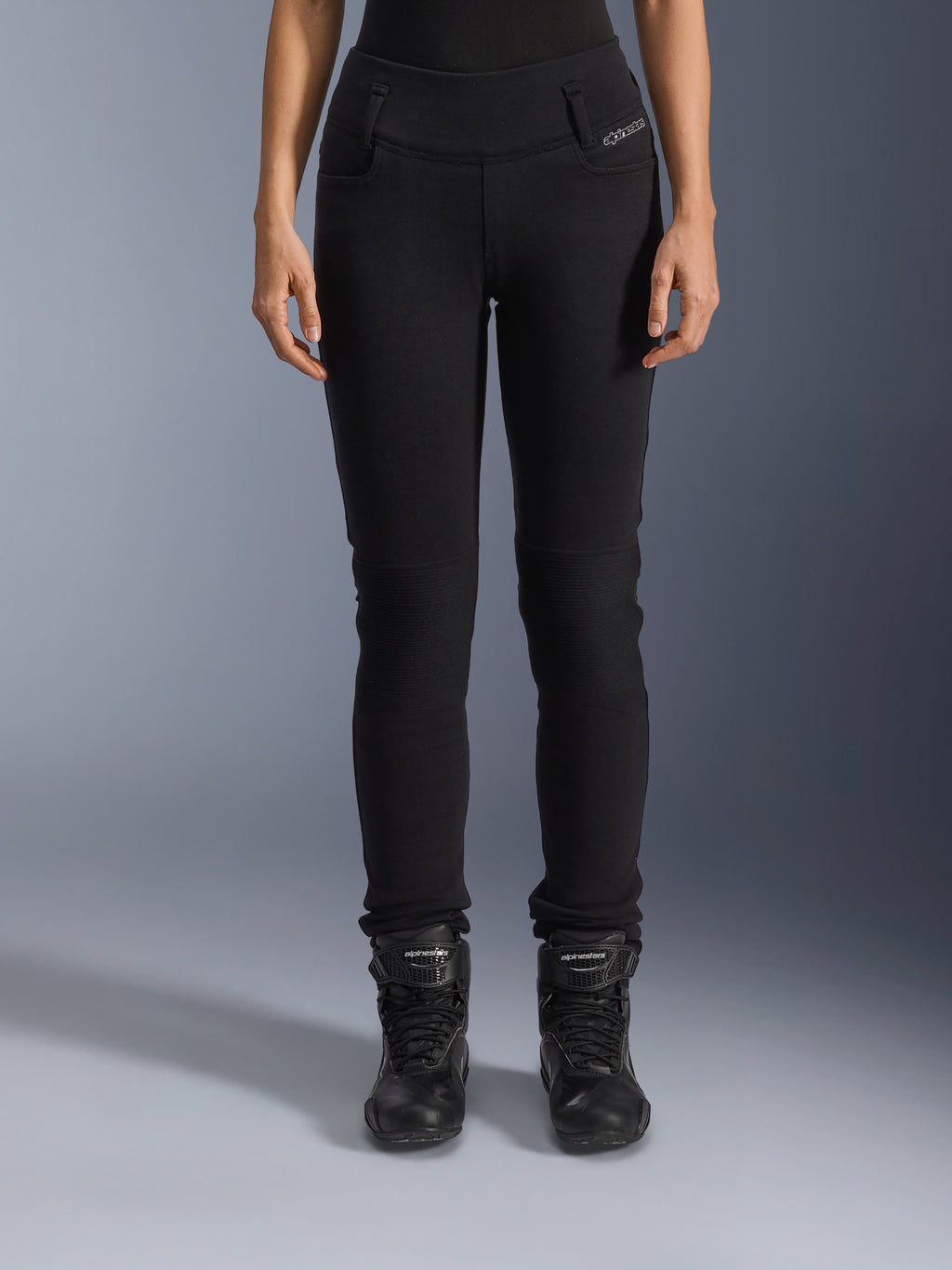Alpinestars Womens Banshee Leggings, czarne spodnie motocyklowe, krój slim-fit z harmonijkowymi panelami na kolanach, noszone z czarnymi wysokimi butami motocyklowymi, miejska odzież motocyklowa w casualowym stylu