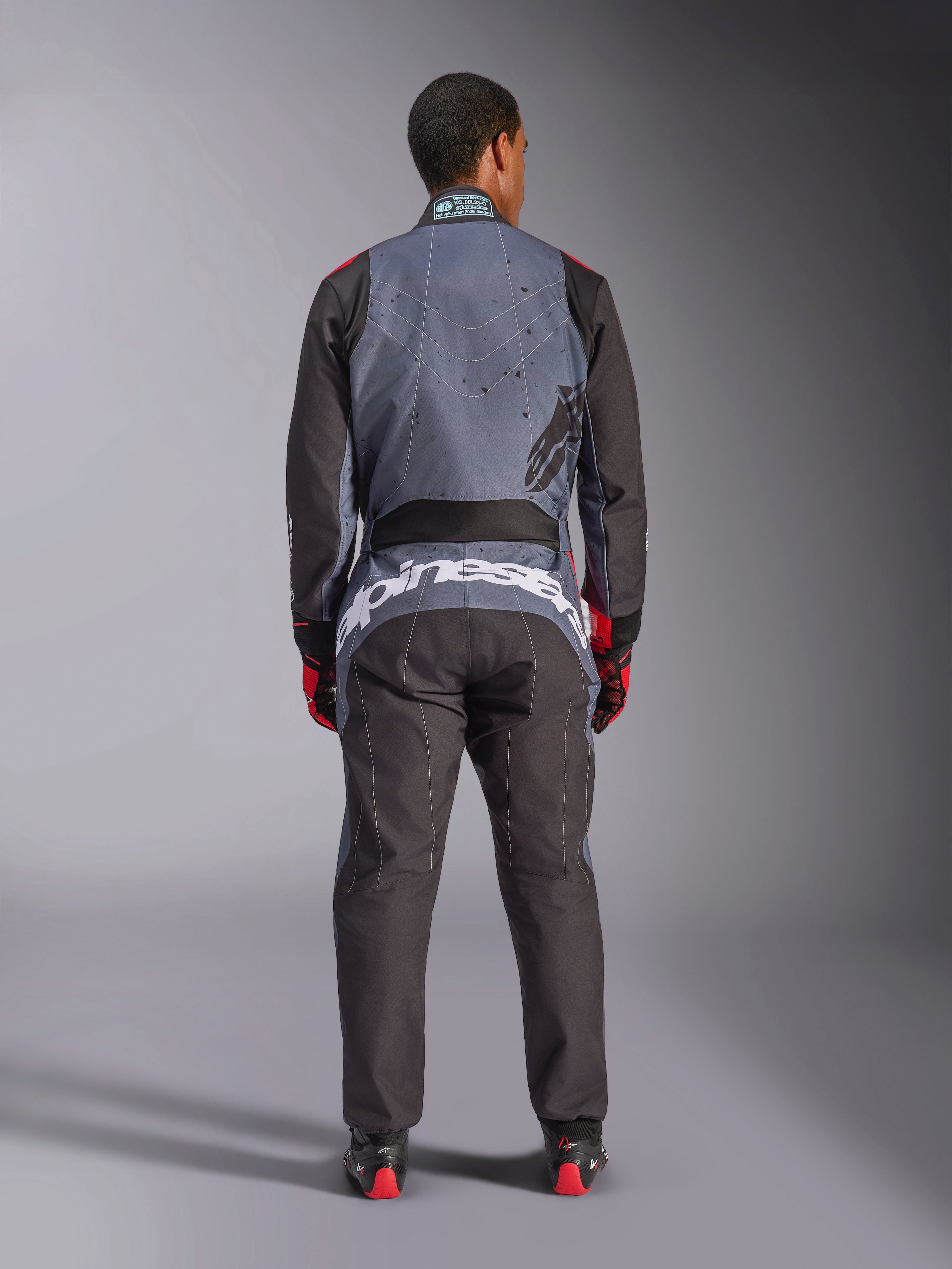 Alpinestars KMX-9 V3 Graphic 6 Suit, kombinezon kartingowy, antracytowa czerń i czerwień, widok z tyłu z dużym białym logo Alpinestars w dolnej części pleców, szara góra z czarną grafiką typu splatter, czarne rękawy i nogawki, noszony z czerwono-czarnymi rękawicami i butami wyścigowymi.