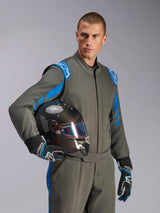 Male driver wearing Alpinestars GP Race V3 Suit FIA in Anthracite Blue, holding a carbon fiber racing helmet with an iridescent visor, professional auto racing gear with blue shoulder accents and matching racing gloves.