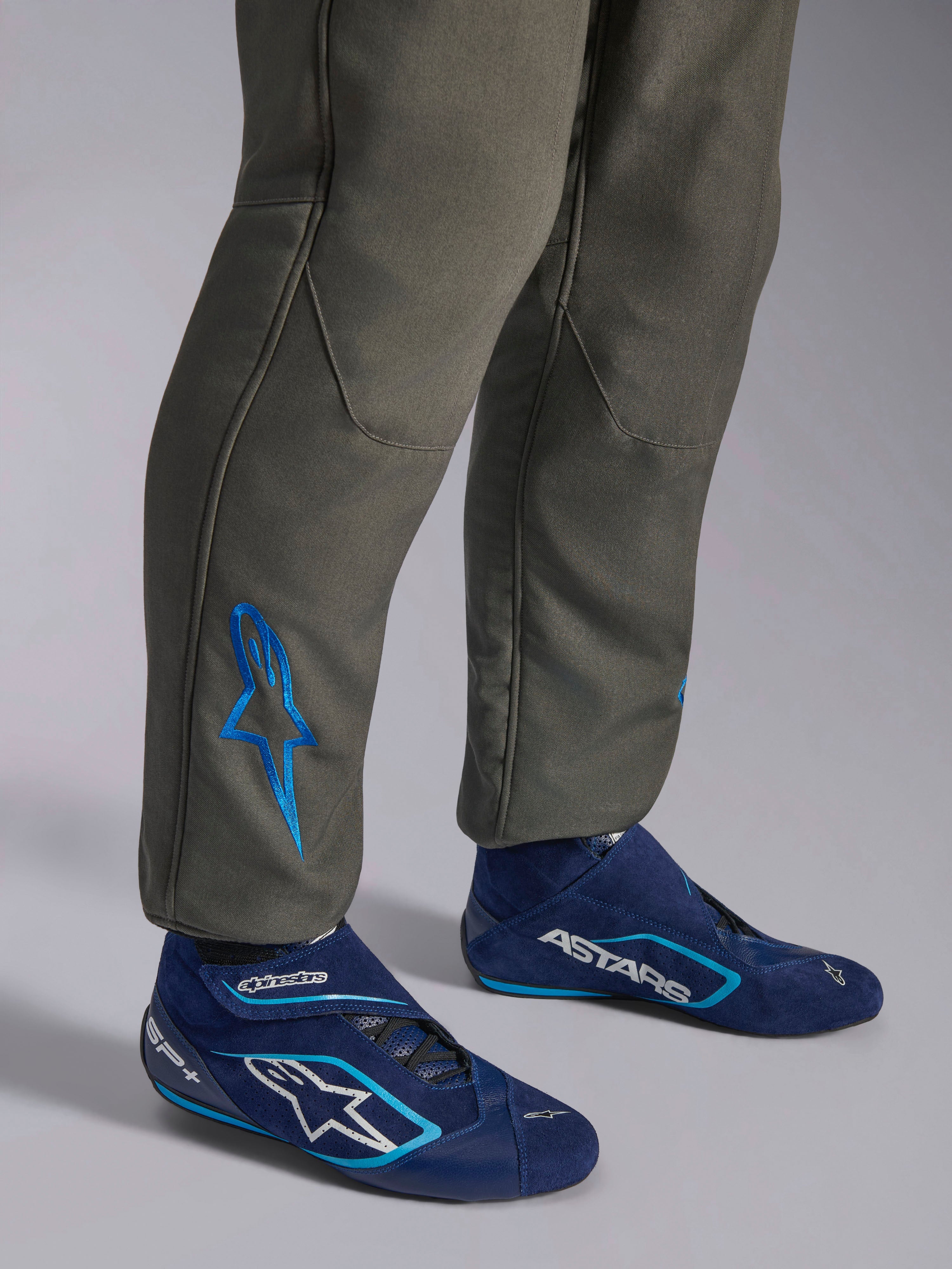 Alpinestars GP Race V3 Suit FIA, kombinezon samochodowy w kolorze Anthracite Blue, zbliżenie na dolną część nogawek z niebieskim haftowanym logo Astars na kostce, granatowe i błękitne buty wyścigowe, profesjonalna odzież ochronna do sportów motorowych