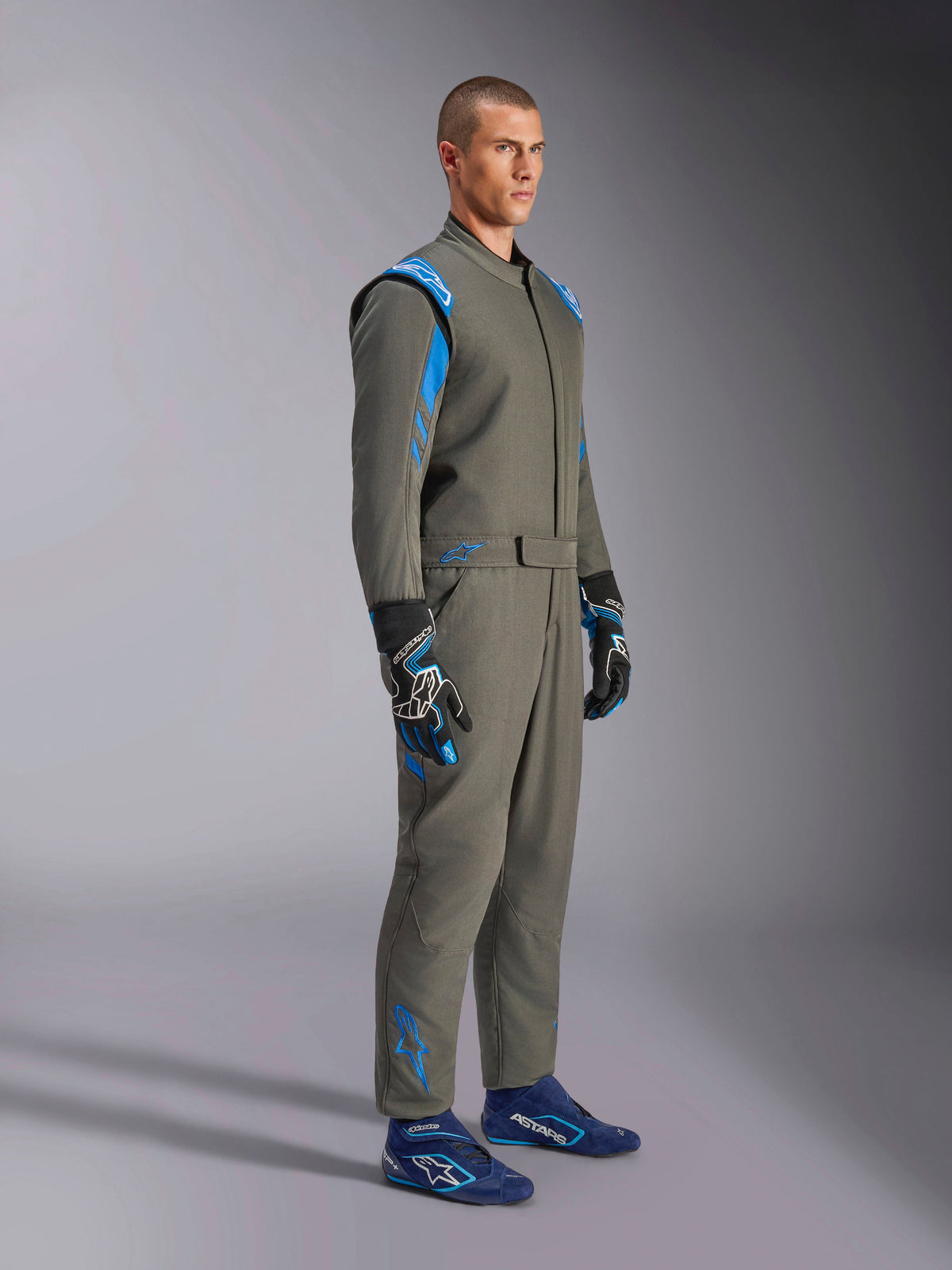 Kombinezon Alpinestars GP Race V3 FIA w kolorze Anthracite Blue, jednoczęściowy samochodowy kombinezon wyścigowy prezentowany na modelu, posiada jasnoniebieskie akcenty na ramionach i kostkach, zestawiony z dopasowanymi rękawicami i niebieskimi butami wyścigowymi, profesjonalny sprzęt do sportów motorowych.