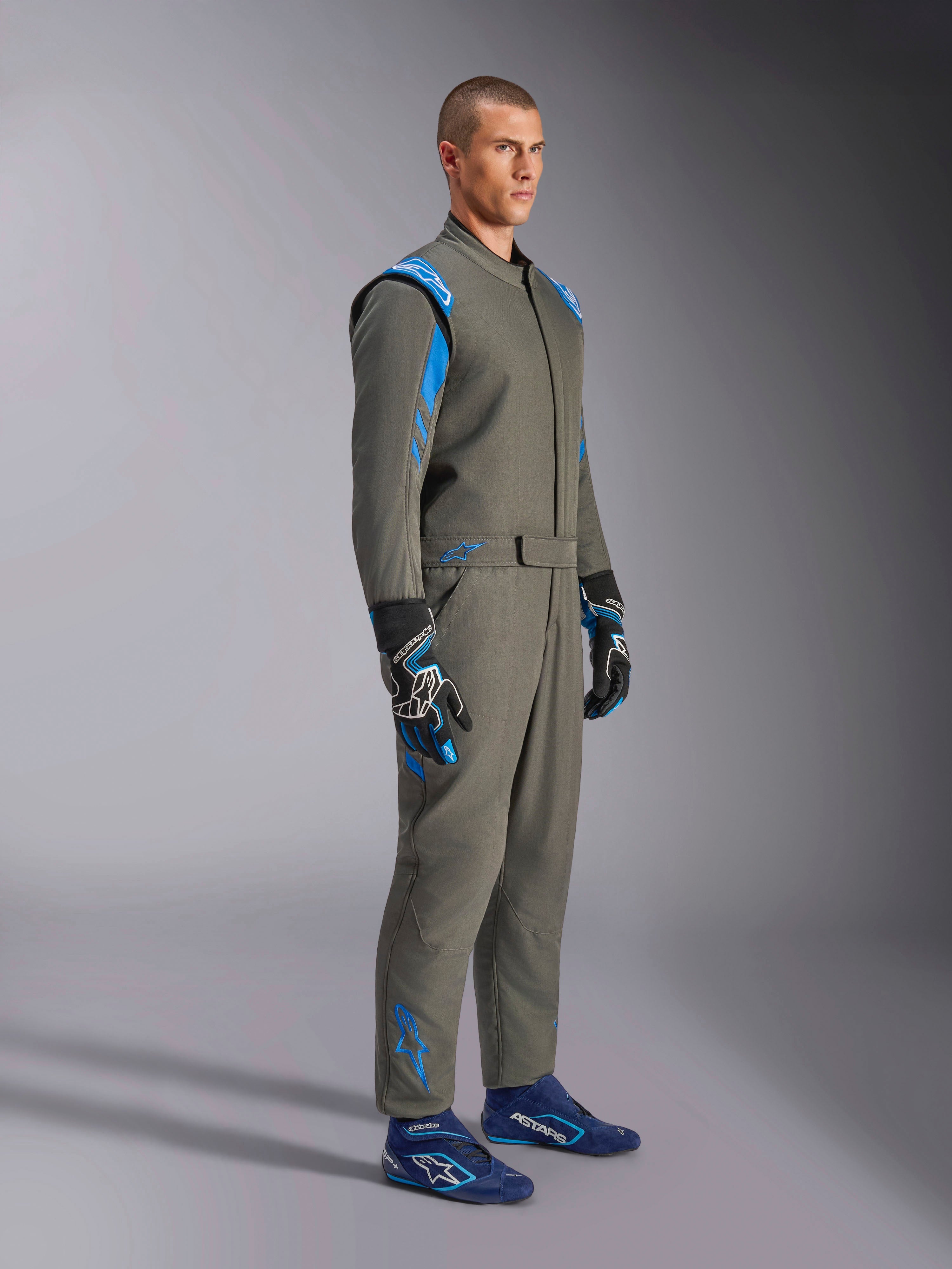 Kombinezon Alpinestars GP Race V3 FIA w kolorze Anthracite Blue, jednoczęściowy samochodowy kombinezon wyścigowy prezentowany na modelu, posiada jasnoniebieskie akcenty na ramionach i kostkach, zestawiony z dopasowanymi rękawicami i niebieskimi butami wyścigowymi, profesjonalny sprzęt do sportów motorowych.