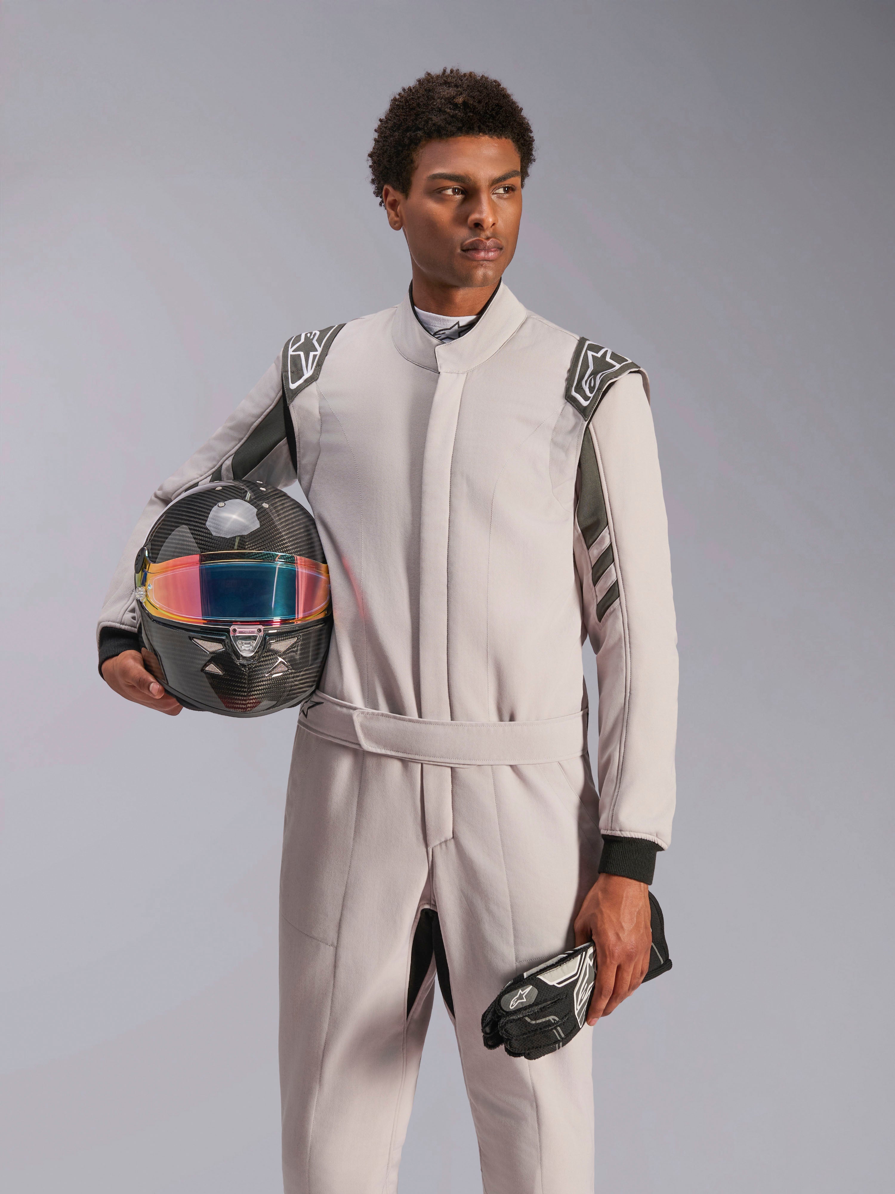 Alpinestars GP Race V3 Suit FIA, kombinezon samochodowy, Silver Anthracite, noszony przez modela trzymającego kask z włókna węglowego z opalizującym wizjerem i czarne rękawice wyścigowe, profesjonalna odzież ochronna do sportów motorowych z widocznymi epoletami na ramionach.