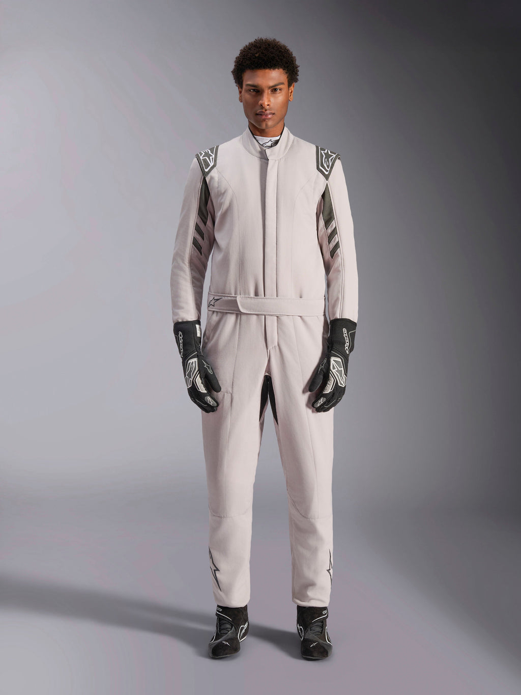 Alpinestars GP Race V3 Suit FIA w kolorze srebrno-antracytowym, jednoczęściowy samochodowy kombinezon wyścigowy prezentowany przez modela, z ciemnoszarymi epoletami i akcentami, czarne rękawice i buty wyścigowe, profesjonalna odzież ochronna do sportów motorowych
