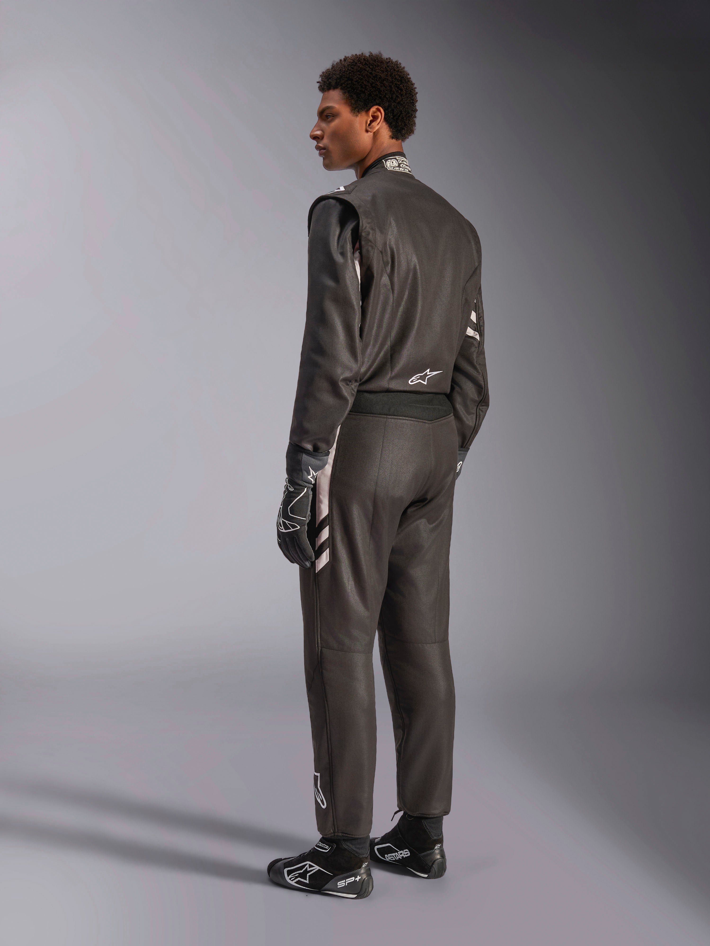 Alpinestars GP Race V3 Suit FIA, czarny samochodowy kombinezon wyścigowy ze srebrnymi akcentami, prezentowany przez modela w widoku z trzech czwartych od tyłu, w zestawie z dopasowanymi czarnymi rękawicami i butami wyścigowymi, profesjonalny sprzęt motorsportowy o eleganckiej dwuwarstwowej konstrukcji.