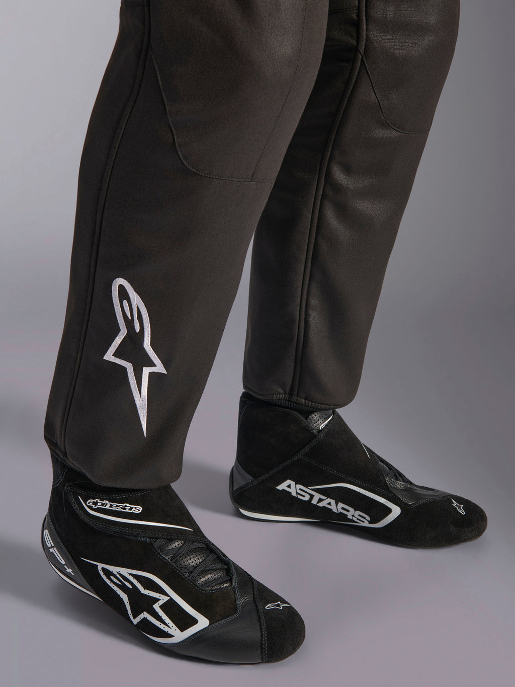 Alpinestars GP Race V3 Suit FIA, zbliżenie na dolną część nogawek i buty wyścigowe, czarny ze srebrnymi akcentami, białe haftowane logo gwiazdy na goleni, prążkowane mankiety przy kostkach, profesjonalna odzież ochronna do wyścigów samochodowych