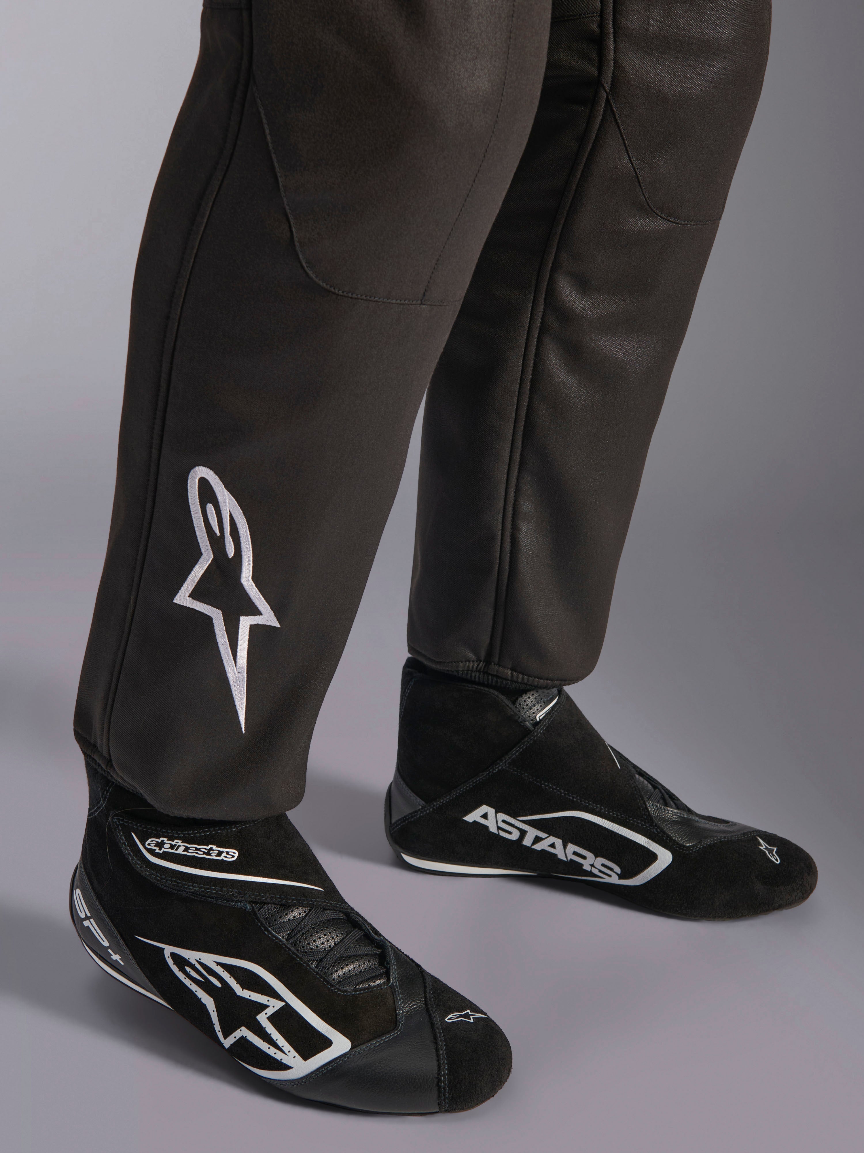 Alpinestars GP Race V3 Suit FIA, zbliżenie na dolną część nogawek i buty wyścigowe, czarny ze srebrnymi akcentami, białe haftowane logo gwiazdy na goleni, prążkowane mankiety przy kostkach, profesjonalna odzież ochronna do wyścigów samochodowych