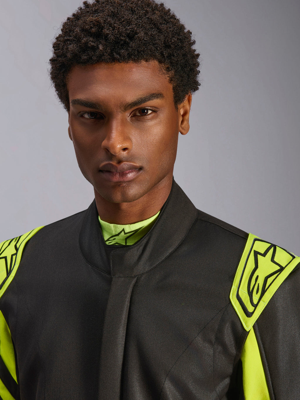 Zbliżenie na modela w kombinezonie Alpinestars GP Race V3 FIA w kolorze czarnym i żółtym neonowym, żółte epolety ewakuacyjne na ramionach z logo gwiazdy, wysoka stójka, profesjonalny strój wyścigowy