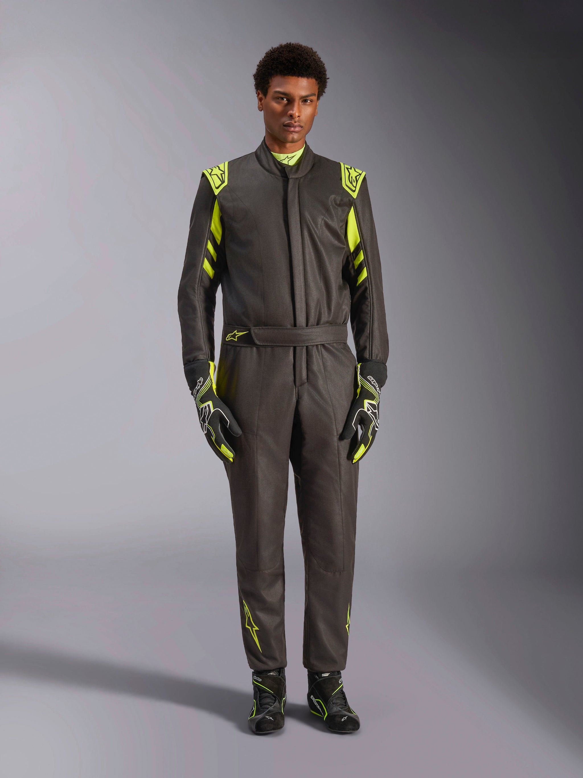 Alpinestars GP Race V3 Suit FIA, kombinezon samochodowy, czarny z neonowymi żółtymi akcentami, prezentowany przez modela z dopasowanymi rękawicami i butami wyścigowymi, jaskrawe detale na ramionach, w pasie i na rękawach, profesjonalny sprzęt motorsportowy