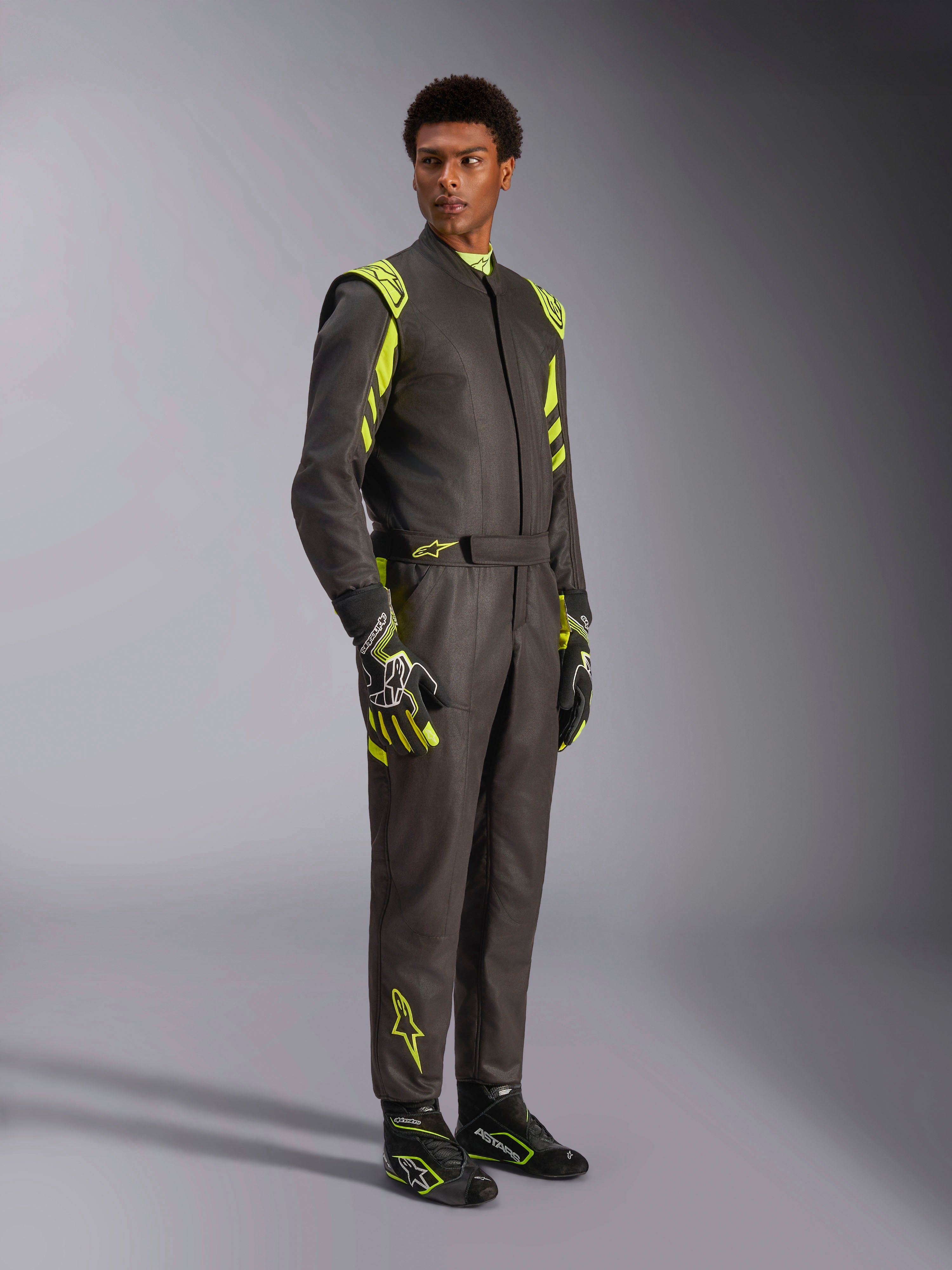 Alpinestars GP Race V3 Suit FIA, kombinezon samochodowy, czarno-żółty fluo, prezentowany przez modela z dopasowanymi rękawicami i butami wyścigowymi, jaskrawe żółte akcenty na ramionach i nogawkach, profesjonalna odzież motorsportowa