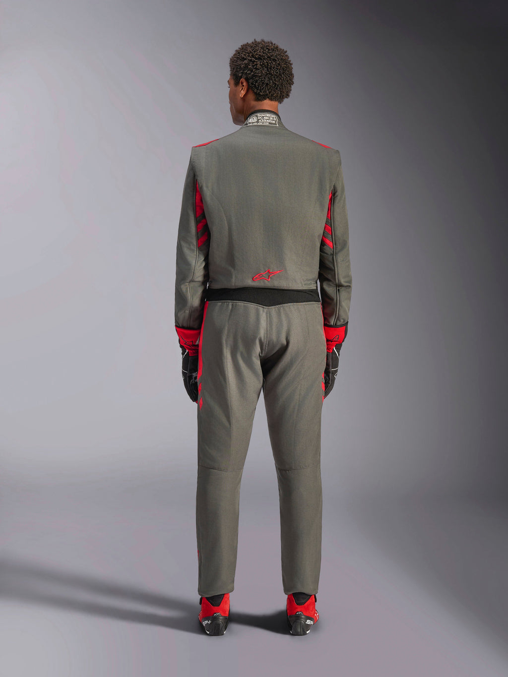 Widok z tyłu modelu ubranego w kombinezon Alpinestars GP Race V3 w kolorze Anthracite Red, profesjonalny kombinezon wyścigowy z czerwonymi akcentami na rękawach i nogawkach, w połączeniu z czerwonymi butami wyścigowymi i czarnymi rękawicami, techniczna odzież motorsportowa pokazana w studio.