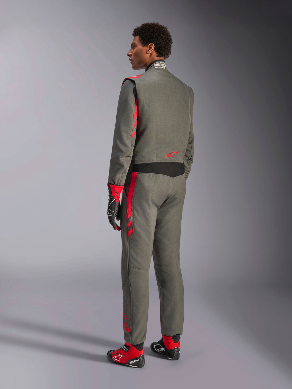 Alpinestars GP Race V3 Suit FIA, antracytowo-czerwony kombinezon wyścigowy, widok od tyłu na modela w dwuwarstwowym kombinezonie, czerwone akcenty na ramionach i nogawkach, dopasowane rękawice i buty, profesjonalna odzież wyścigowa