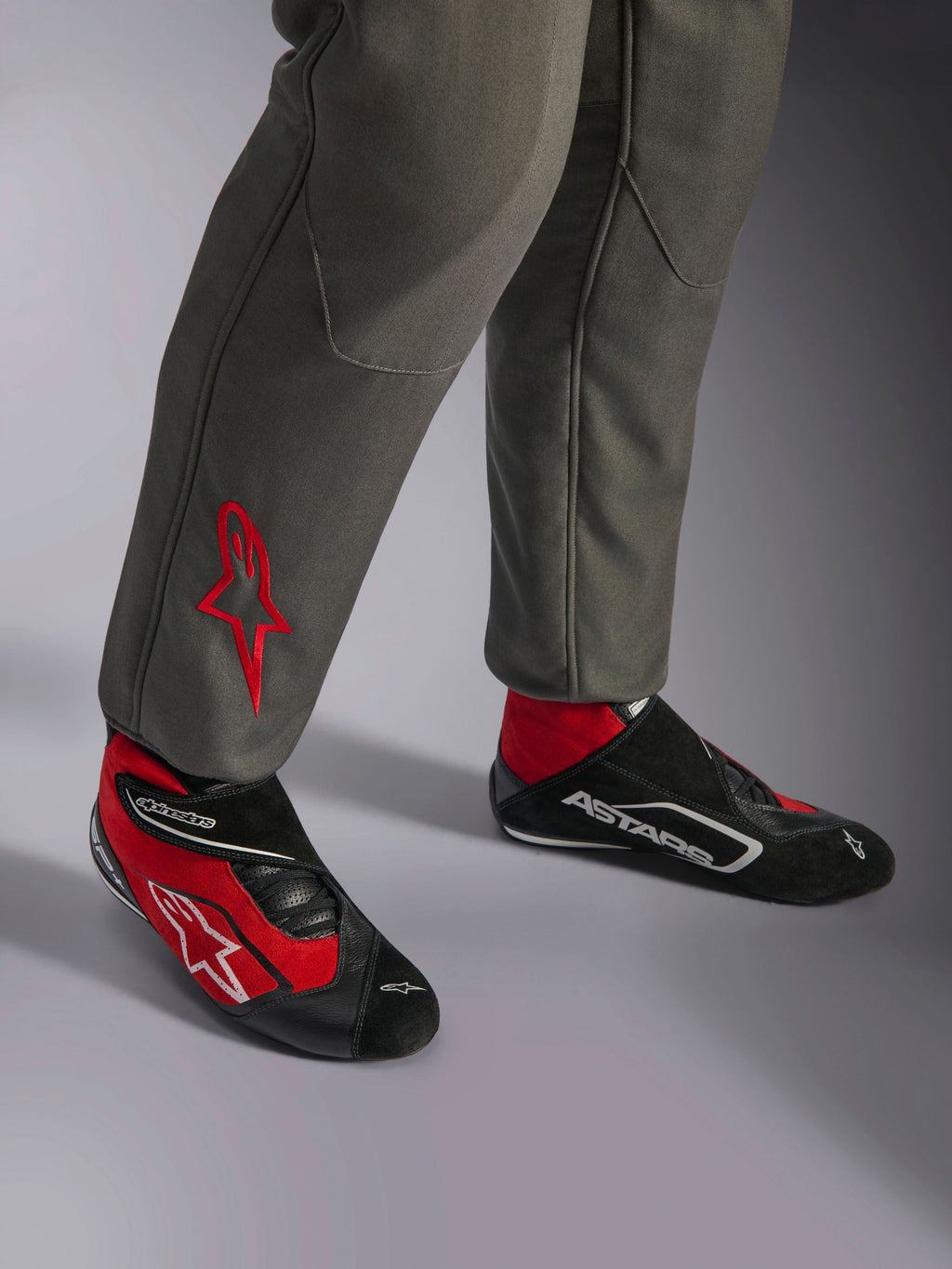 Alpinestars GP Race V3 Suit FIA, kombinezon wyścigowy w kolorze antracytowo-czerwonym, zbliżenie na dolną część nogawek z haftowanym logo czerwonej gwiazdy, w połączeniu z czerwono-czarnymi butami wyścigowymi, profesjonalny sprzęt samochodowy zaprojektowany dla ochrony i komfortu kierowcy